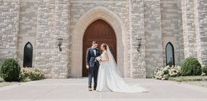 A couple poses in front of a stone church with gothic arched doorway and windows.