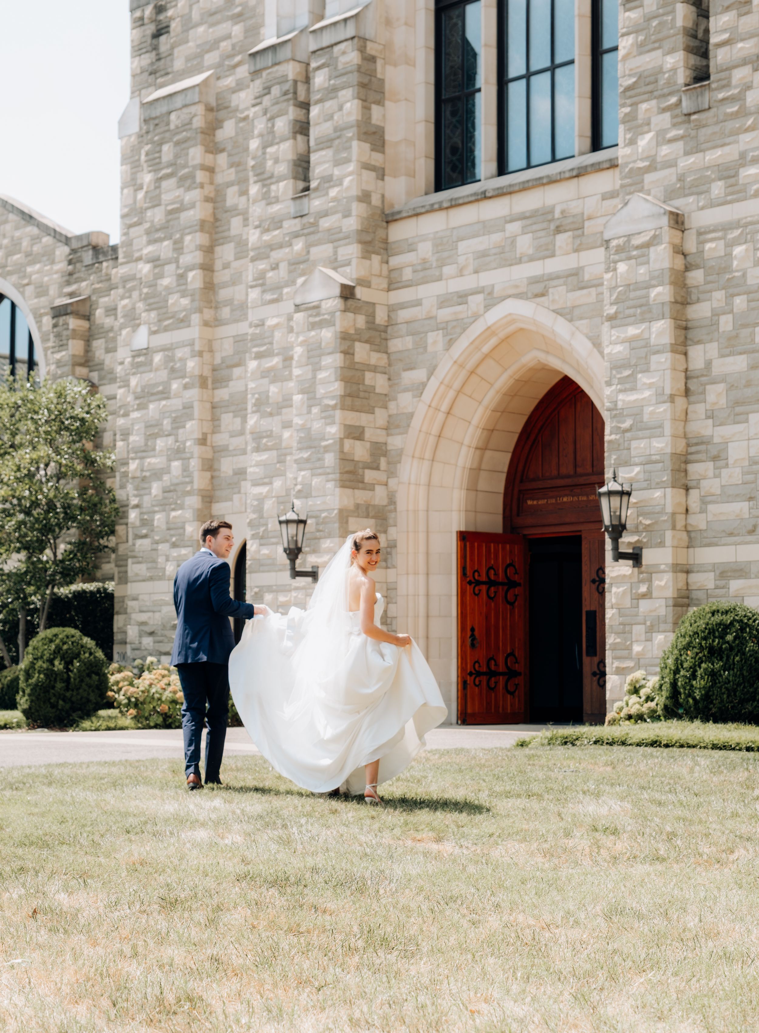 A couple in wedding attire stands on a lawn outside a stone church with arched doorway and lanterns.