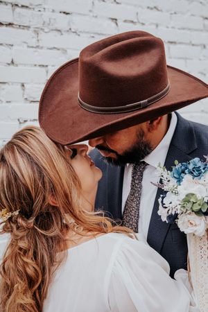 An intimate western wedding photo showing a couple under a brown cowboy hat with blue flowers.