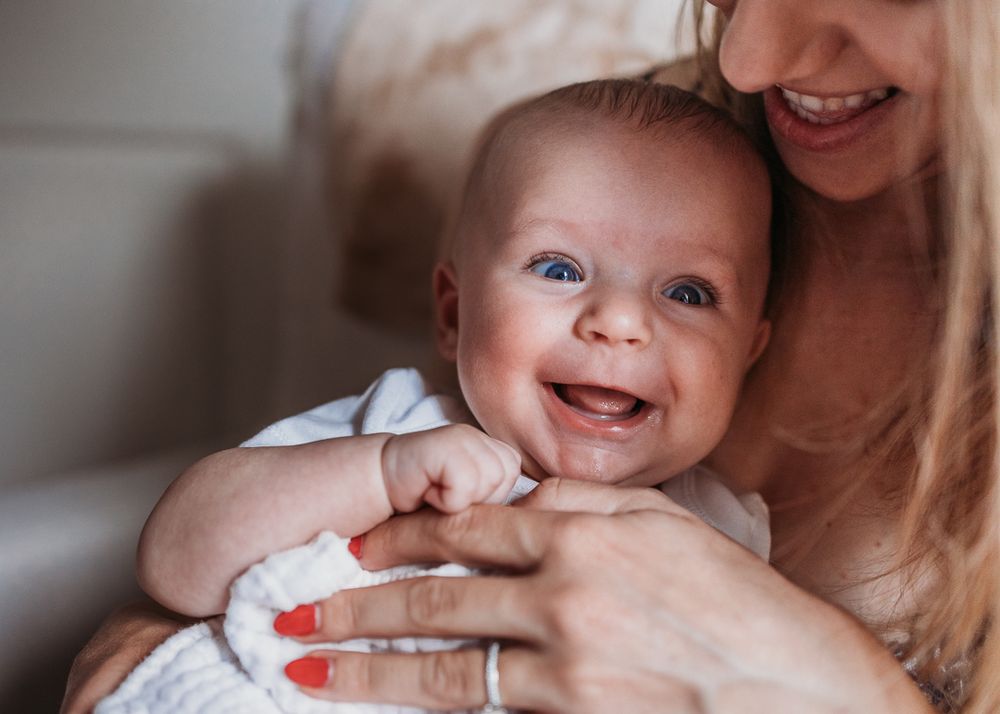 A smiling baby with rosy cheeks happily plays while being held in caring arms with red-painted nails.