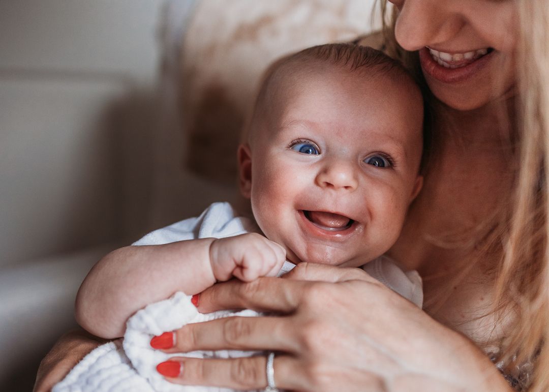 A smiling baby with rosy cheeks happily plays while being held in caring arms with red-painted nails.