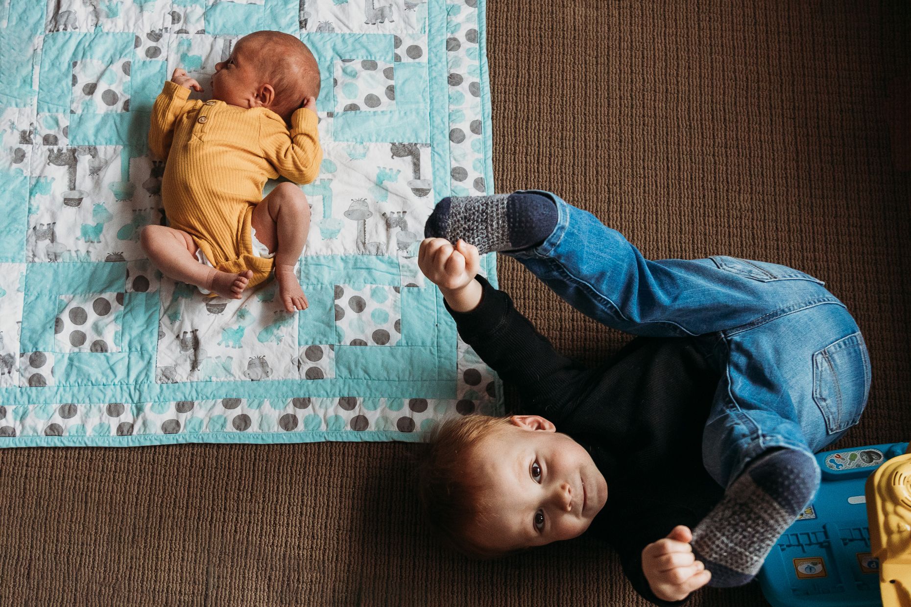 Two children lie on a brown floor with a turquoise patterned blanket, one in yellow and one in blue denim.