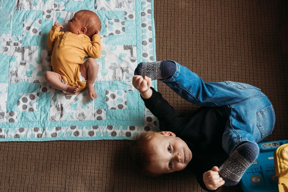 Two children lie on a brown floor with a turquoise patterned blanket, one in yellow and one in blue denim.