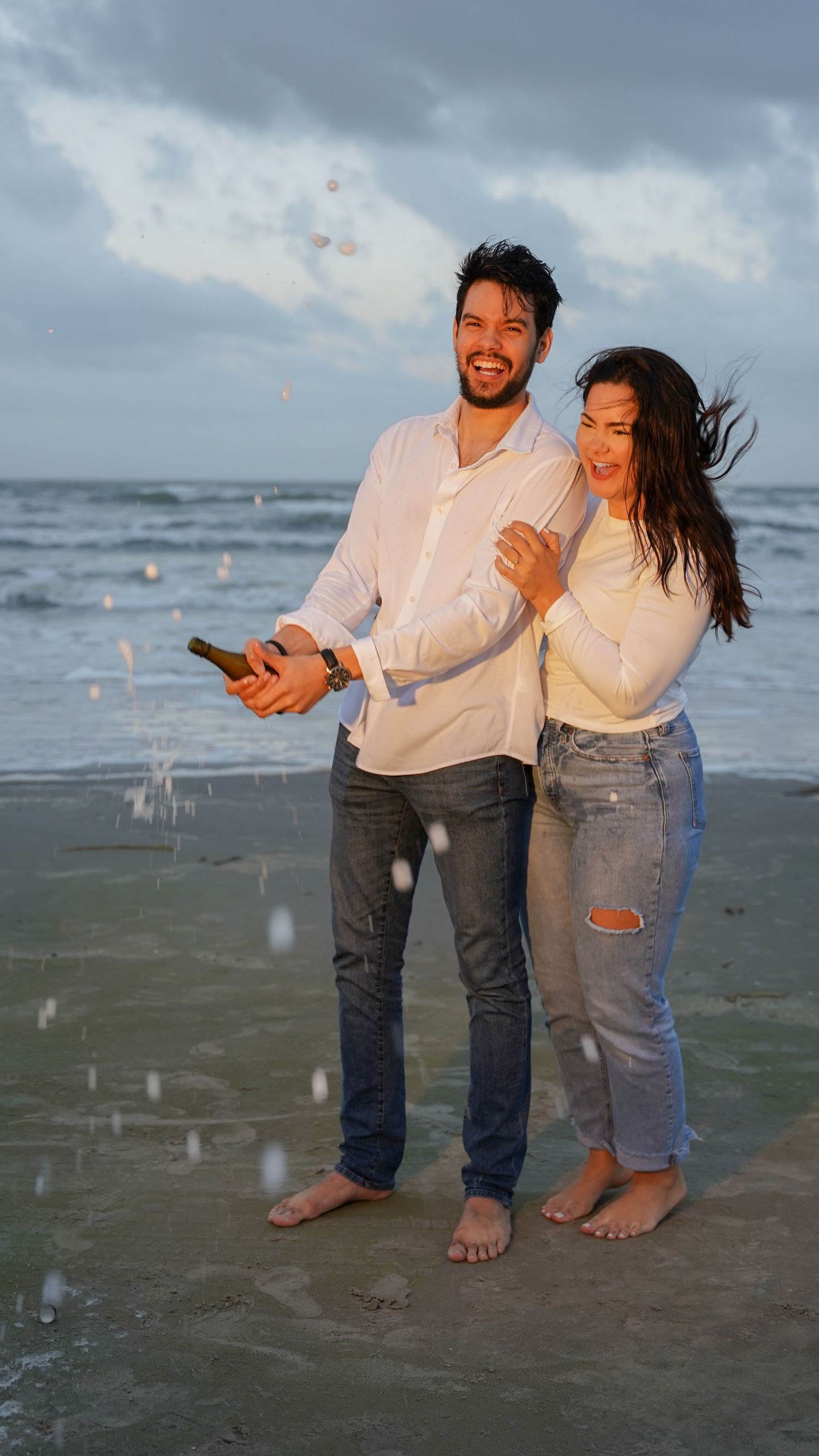 Two people in casual attire share a joyful moment while standing barefoot on a beach at sunset as confetti falls around them.