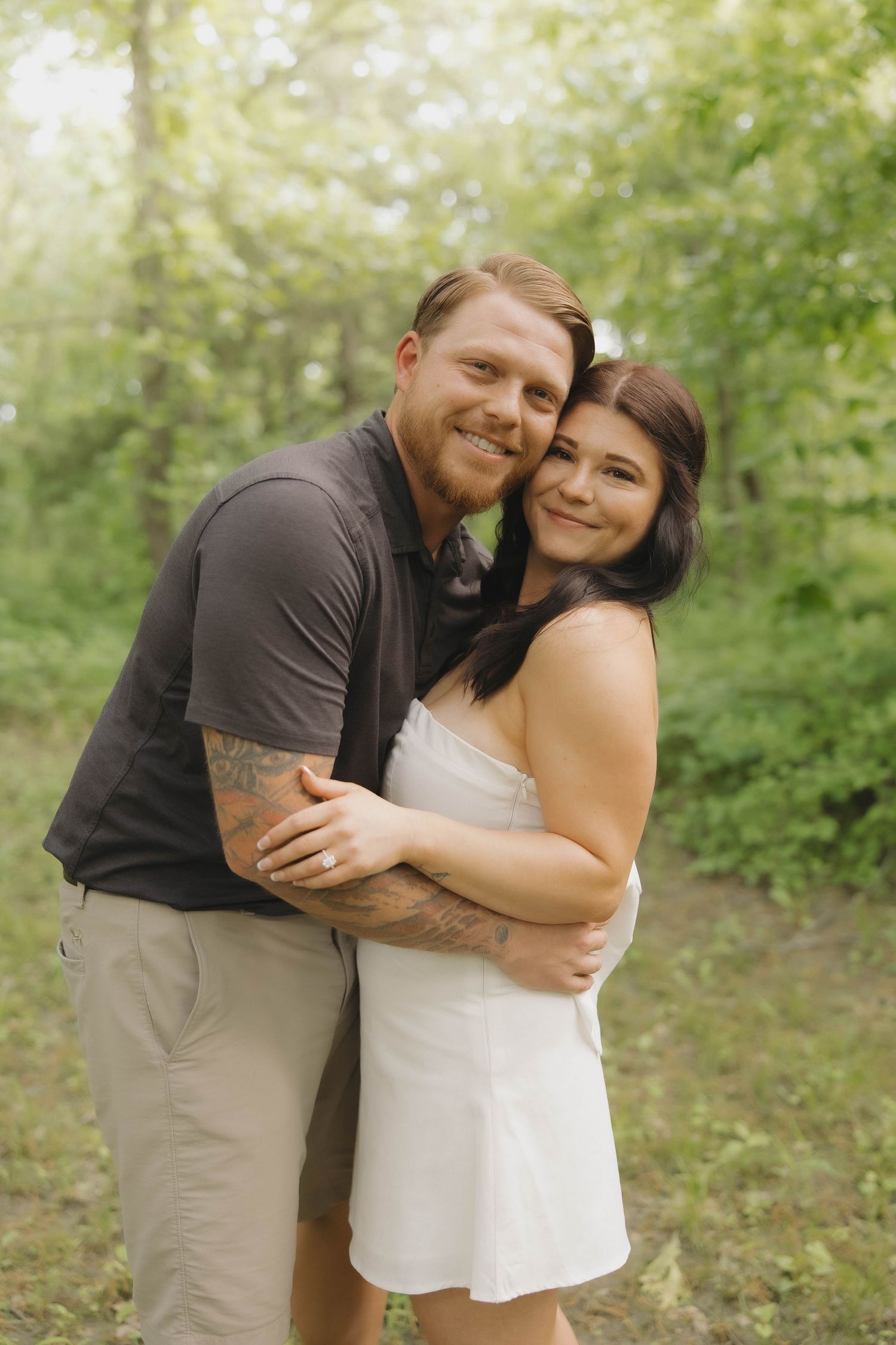 A loving couple embraces and smiles while standing together on a wooded path in natural lighting.