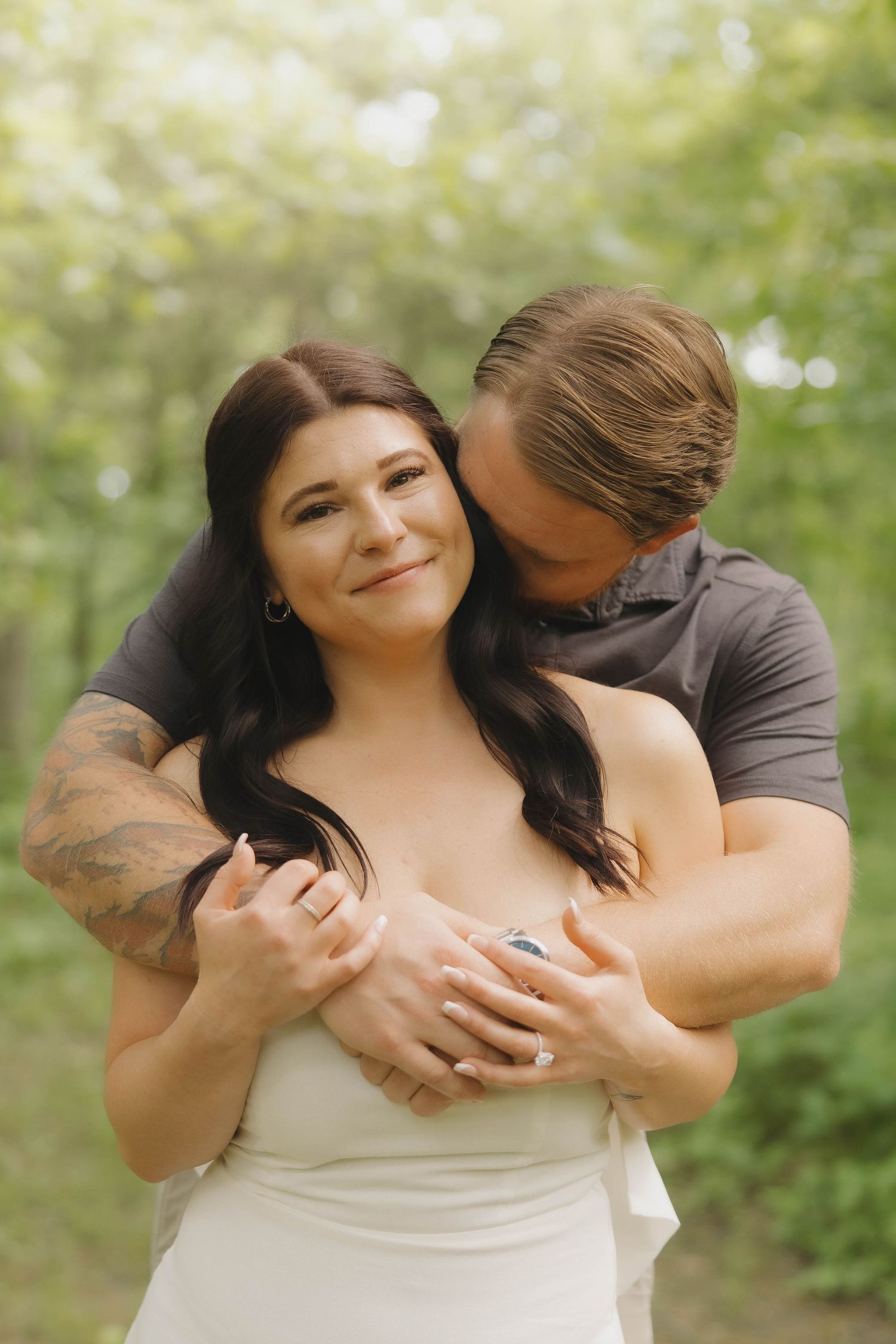 A couple shares an intimate embrace outdoors with natural bokeh background during an engagement photo session.