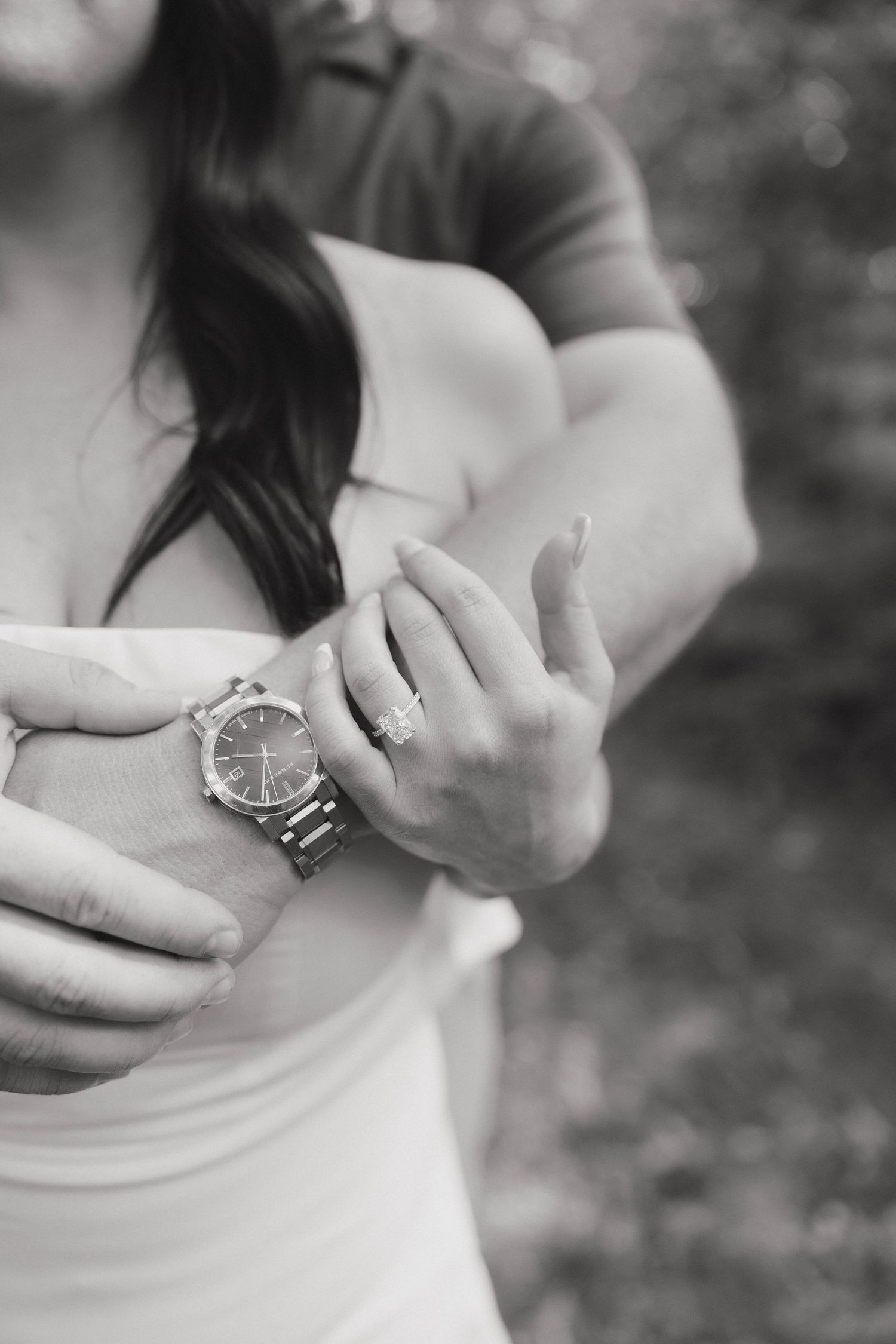 Black and white close-up photo showing hands adjusting a silver wristwatch with ring on finger against blurred background.