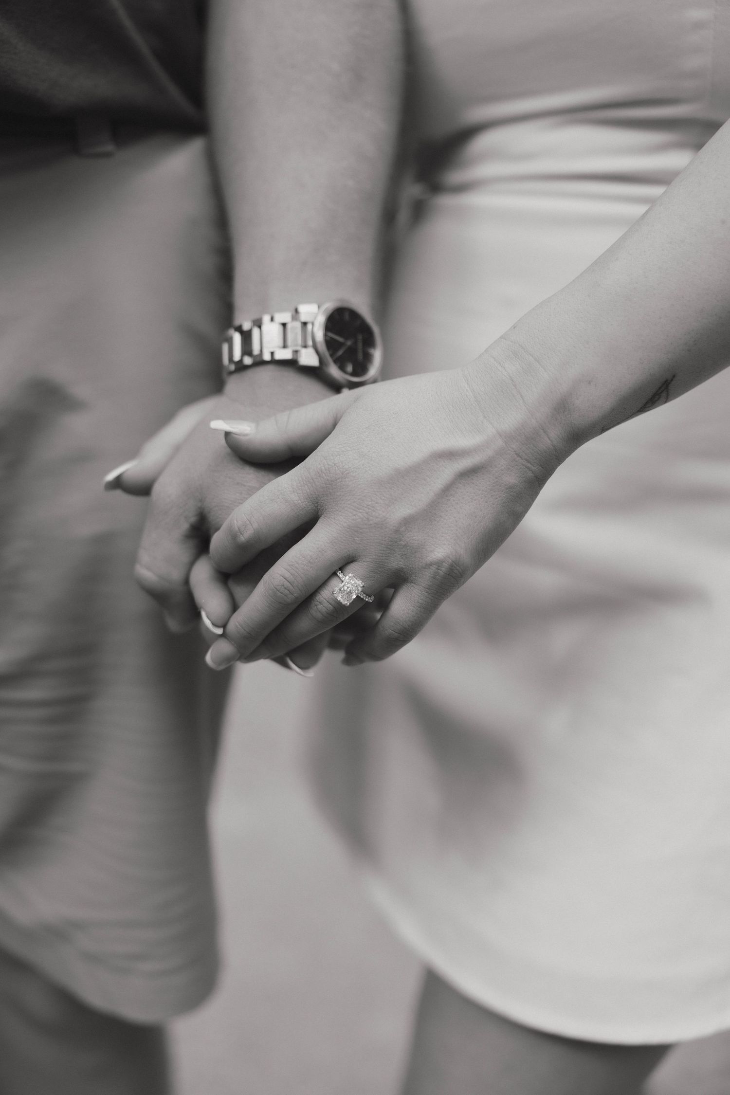 Black and white closeup photo of clasped hands showing a shiny engagement ring and mens watch.
