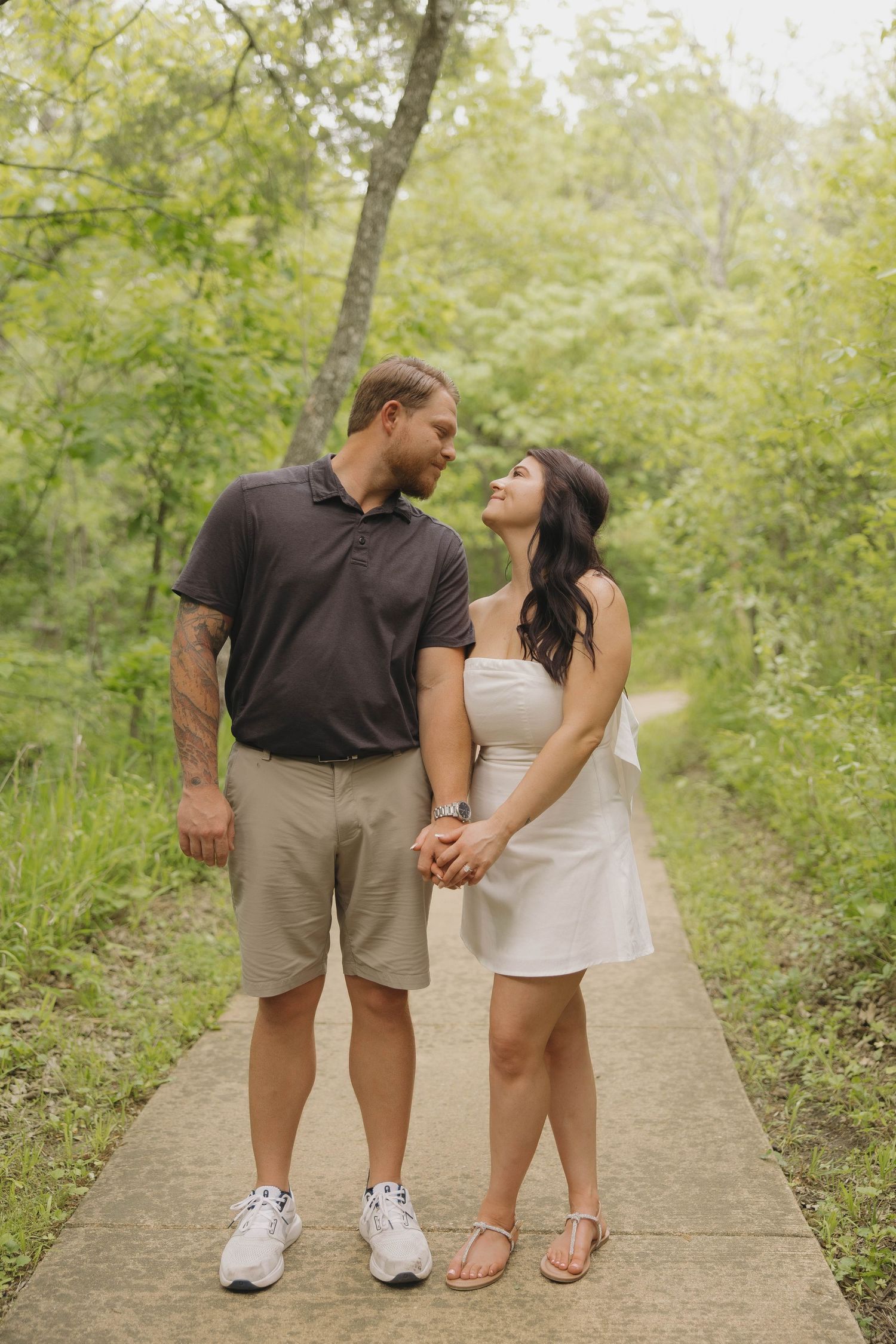 A couple in casual summer attire share a romantic moment while standing on a paved path surrounded by lush greenery.