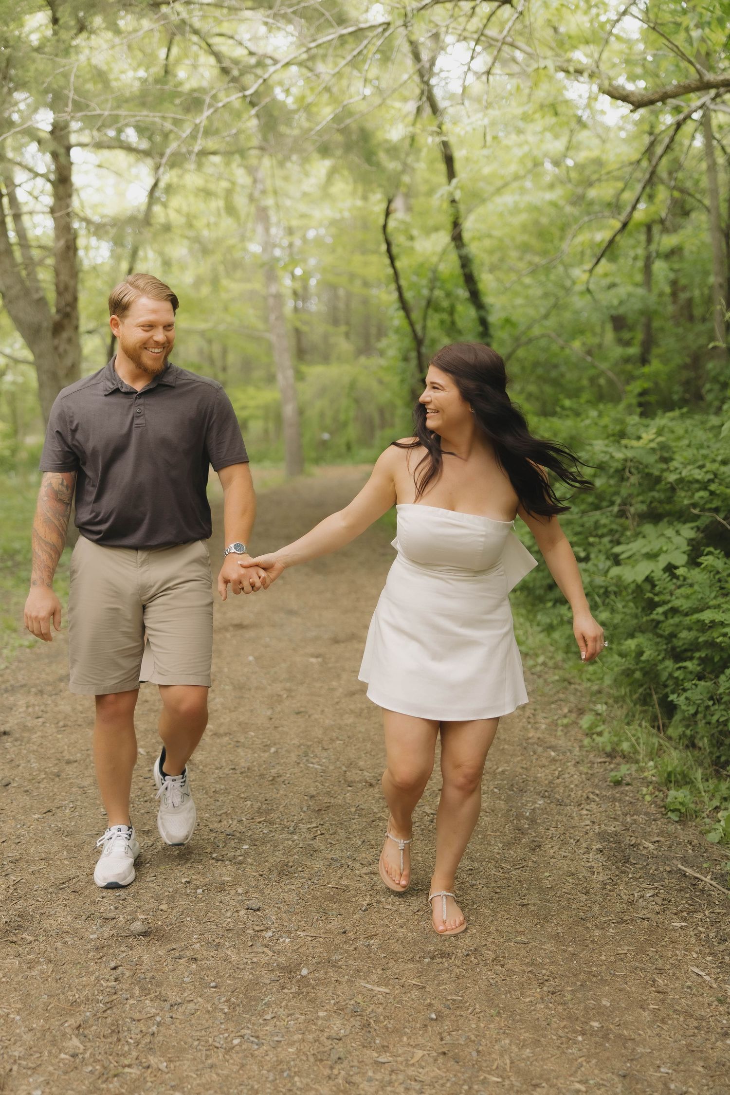 A couple walking hand in hand on a forest trail during summer, with lush green foliage surrounding them.