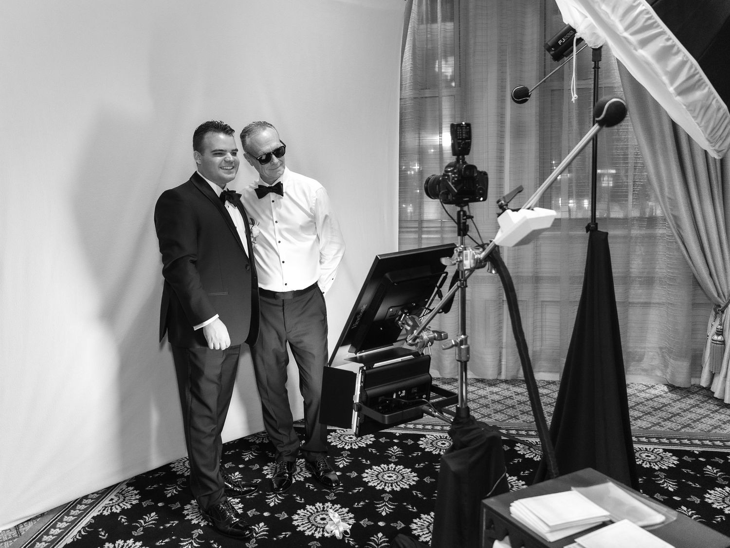 A black and white studio photo booth setup shows professional lighting equipment, umbrella reflector and a couple posing for a portrait