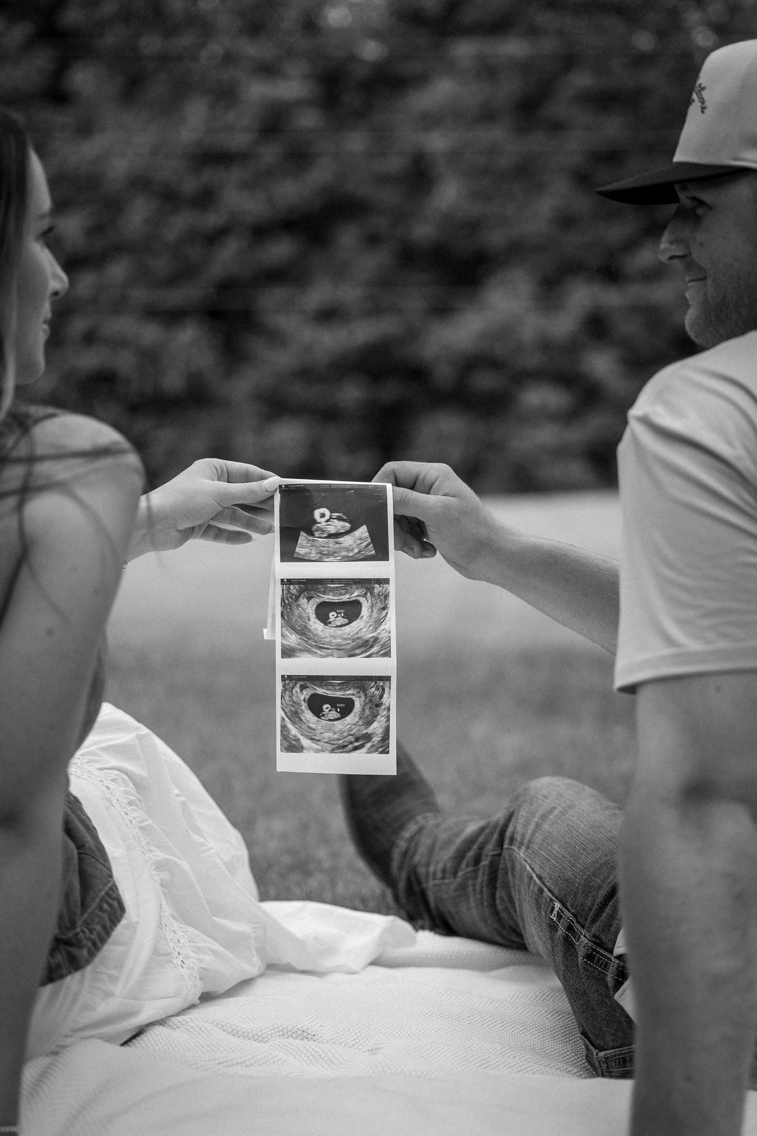 A couple sits outdoors holding up ultrasound photos together in a black and white pregnancy announcement photo.