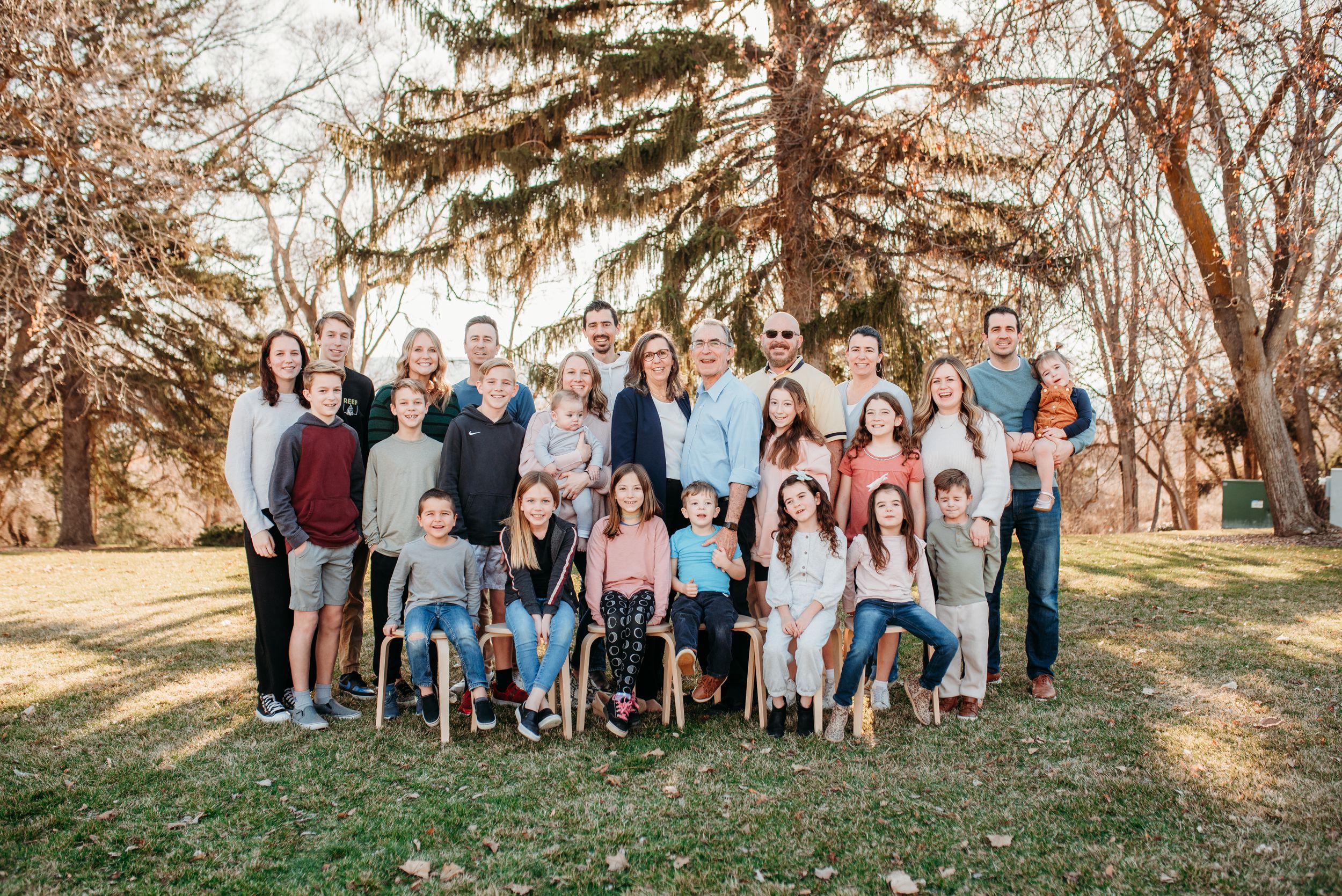 A large extended family group poses together on grass with evergreen trees in the background during autumn.