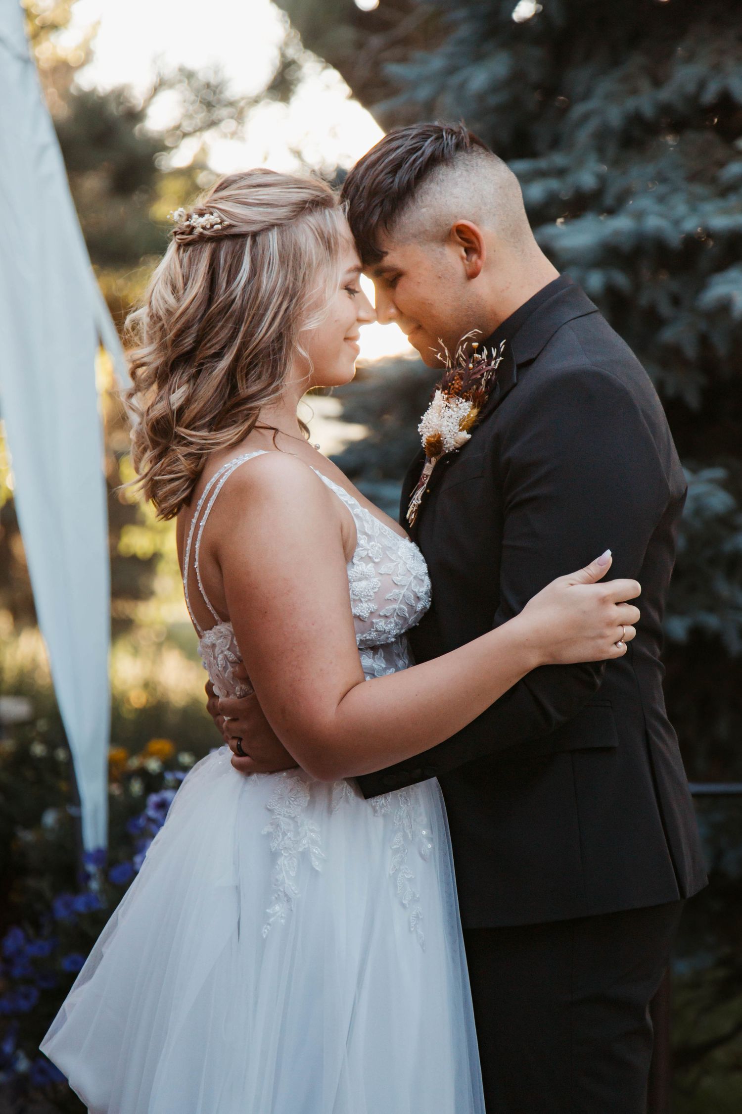 A romantic wedding moment captured outdoors with the couple embracing against a backdrop of blue spruce trees at sunset.