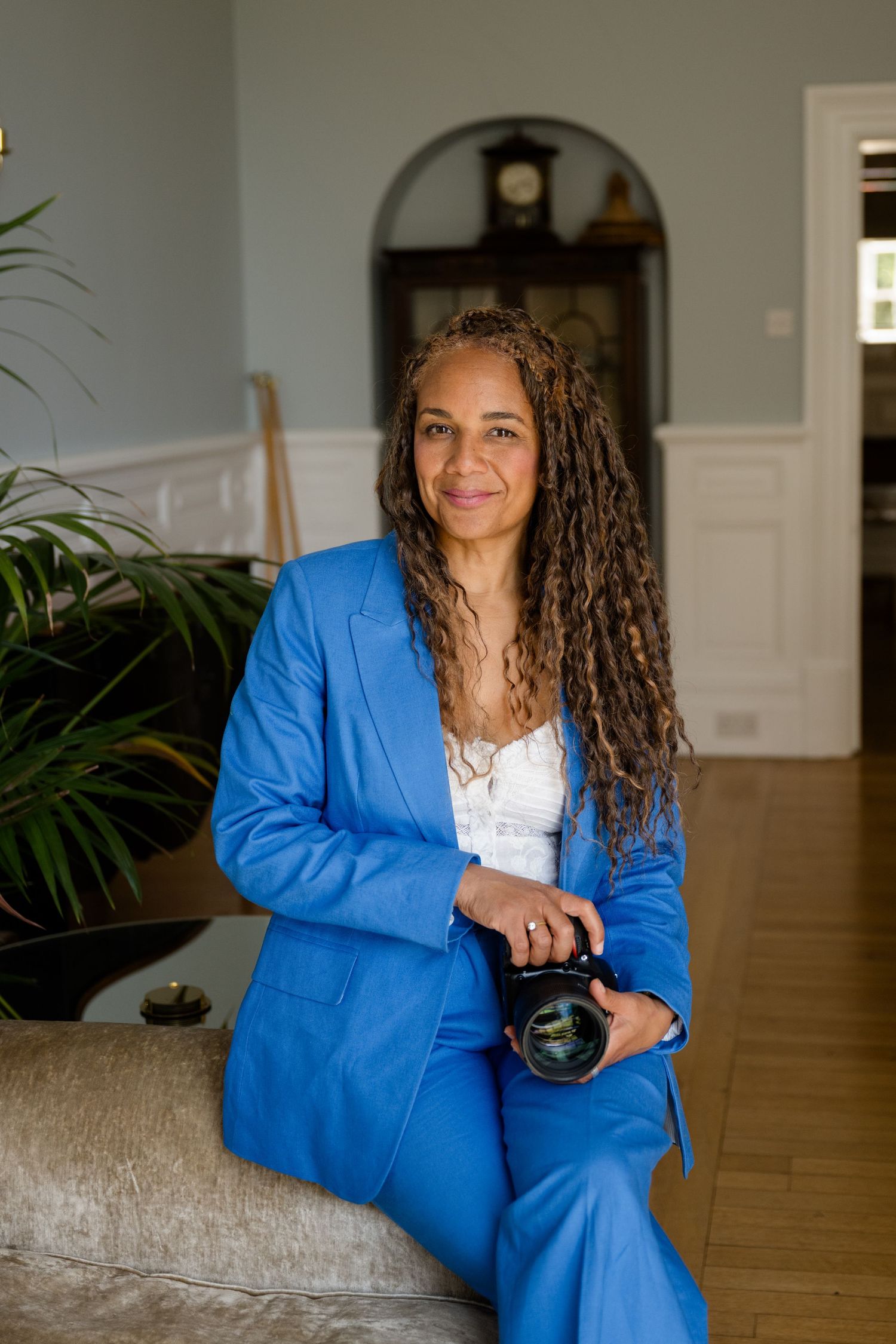 Portrait of a brand photographer on a brand shoot in a location home wearing a white top and blue suit against neutral background.