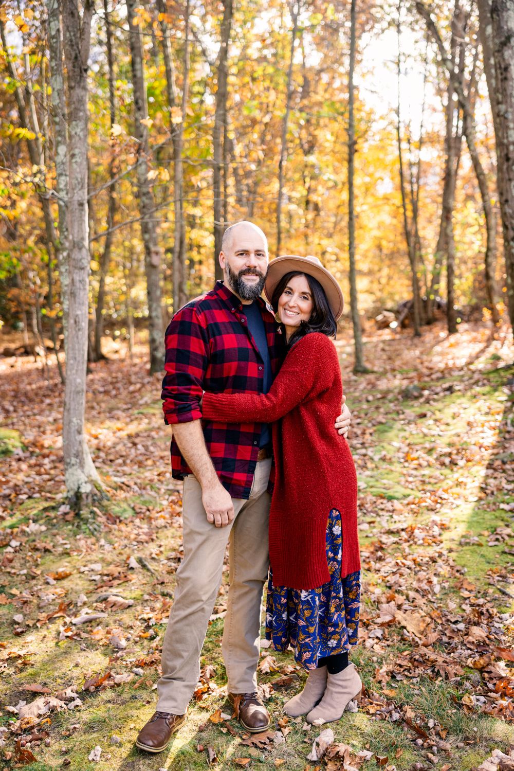 Couple in casual fall attire embracing and smiling in a wooded setting with autumn leaves.