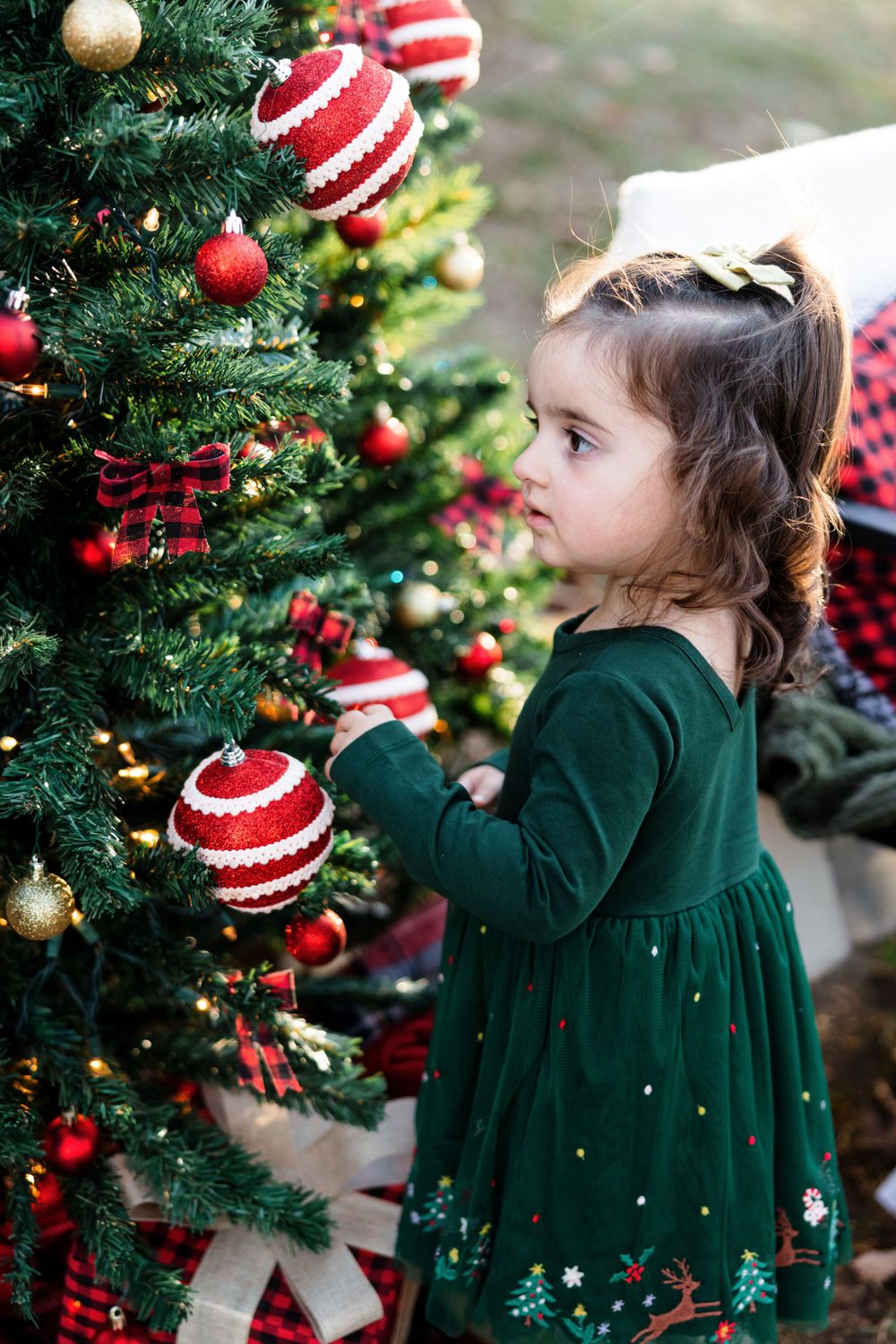 Small child in emerald green dress explores decorated Christmas tree with red and white ornaments.