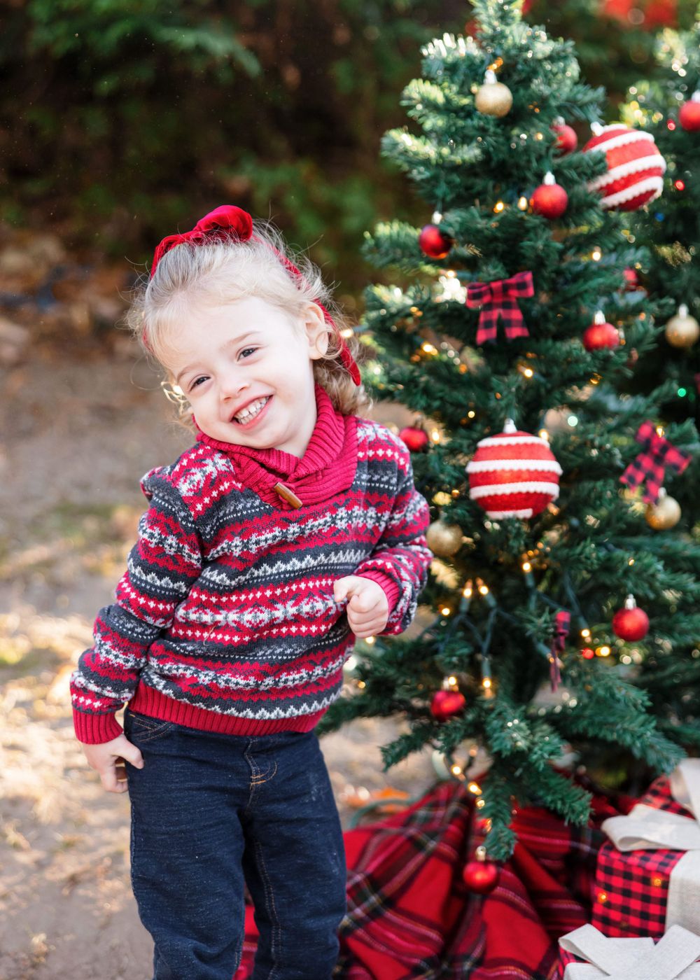 Child wearing festive red and navy fair isle sweater laughs by decorated Christmas tree.