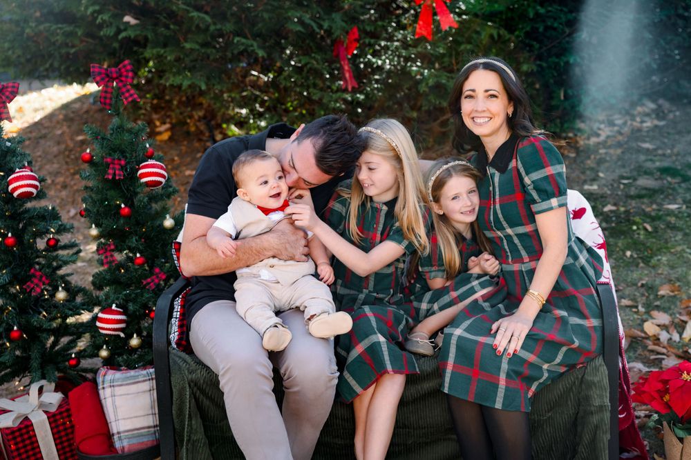 Family wearing matching plaid outfits poses together outdoors near red flowers.