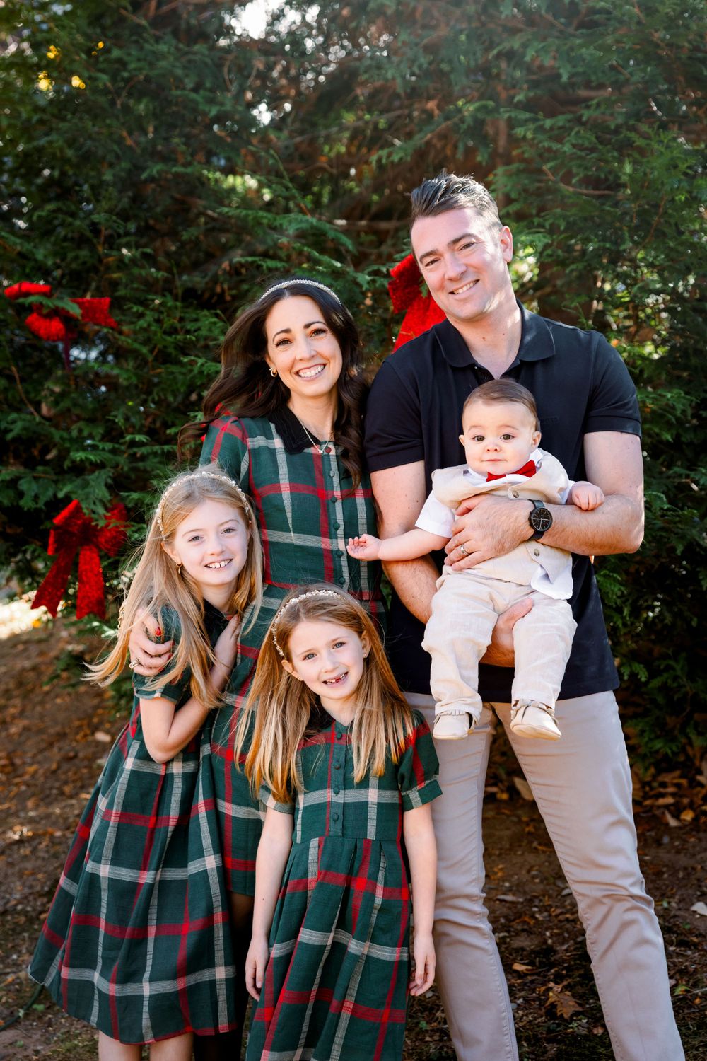 Family wearing coordinated green plaid and black outfits pose together for holiday photos in front of evergreen trees.