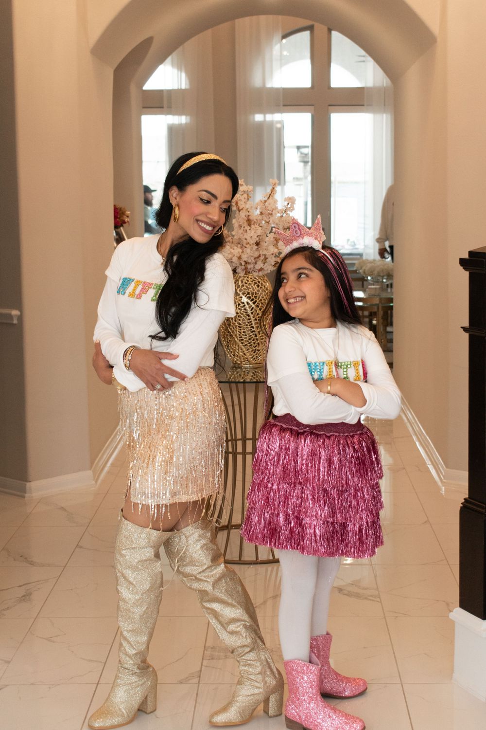 Two people in matching white shirts and sparkly skirts posing together in an elegant entryway.