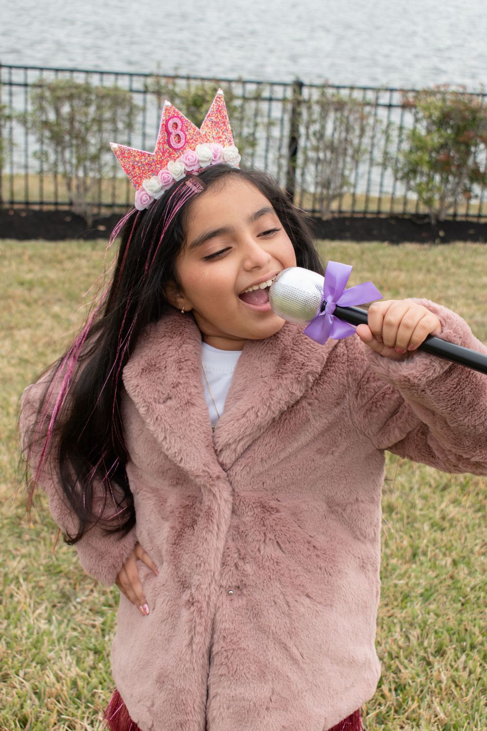 Person wearing a pink crown and fuzzy jacket sings into a microphone while celebrating outdoors.