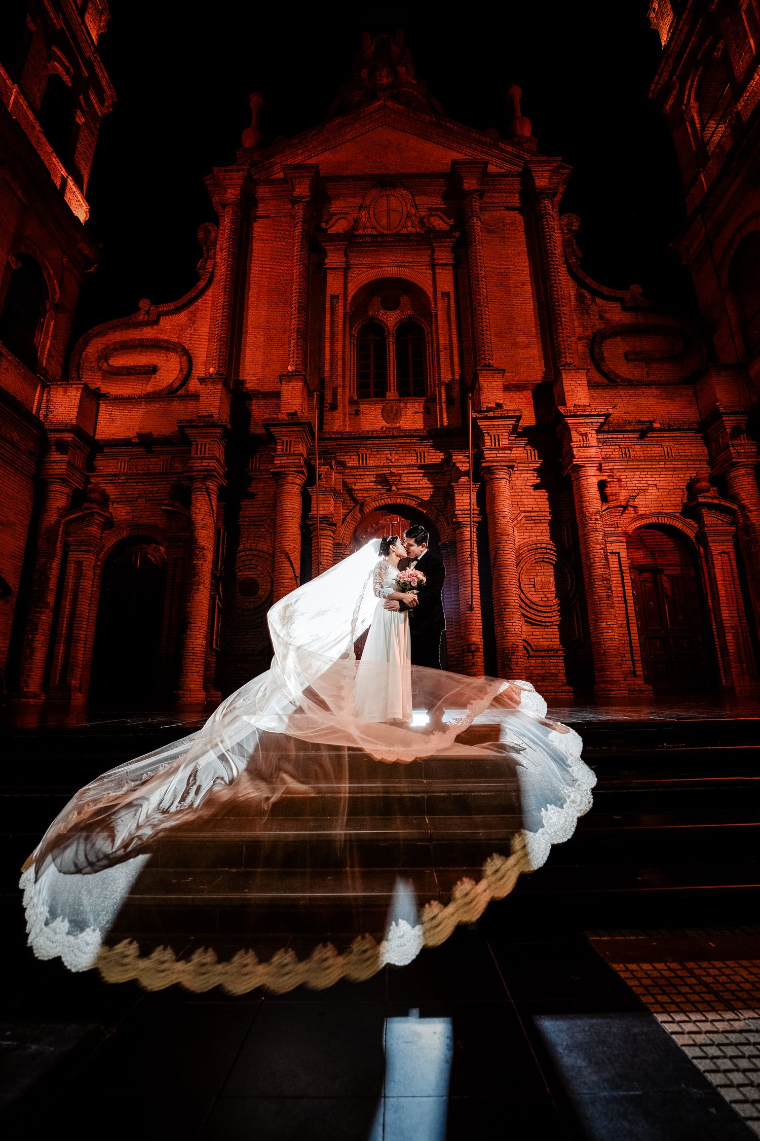 Bride and groom embrace in front of grand, illuminated historic building, with the bride’s flowing veil creating a dramatic, ethereal effect