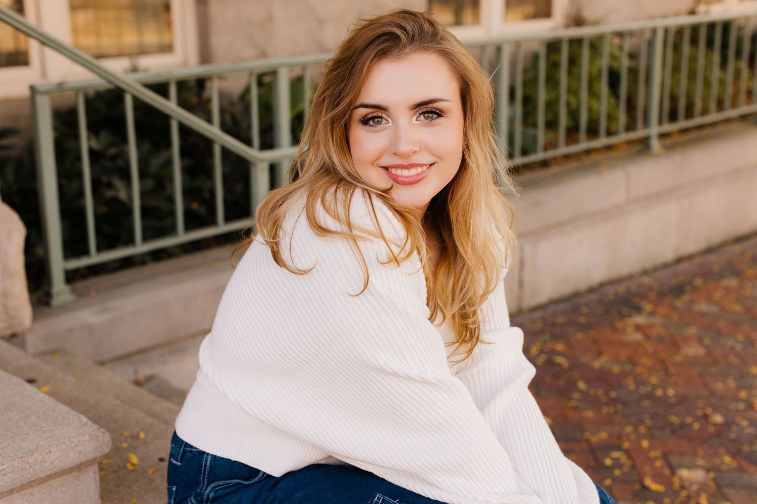 Someone sitting on steps wearing a cozy white sweater and blue jeans while smiling on an autumn day.