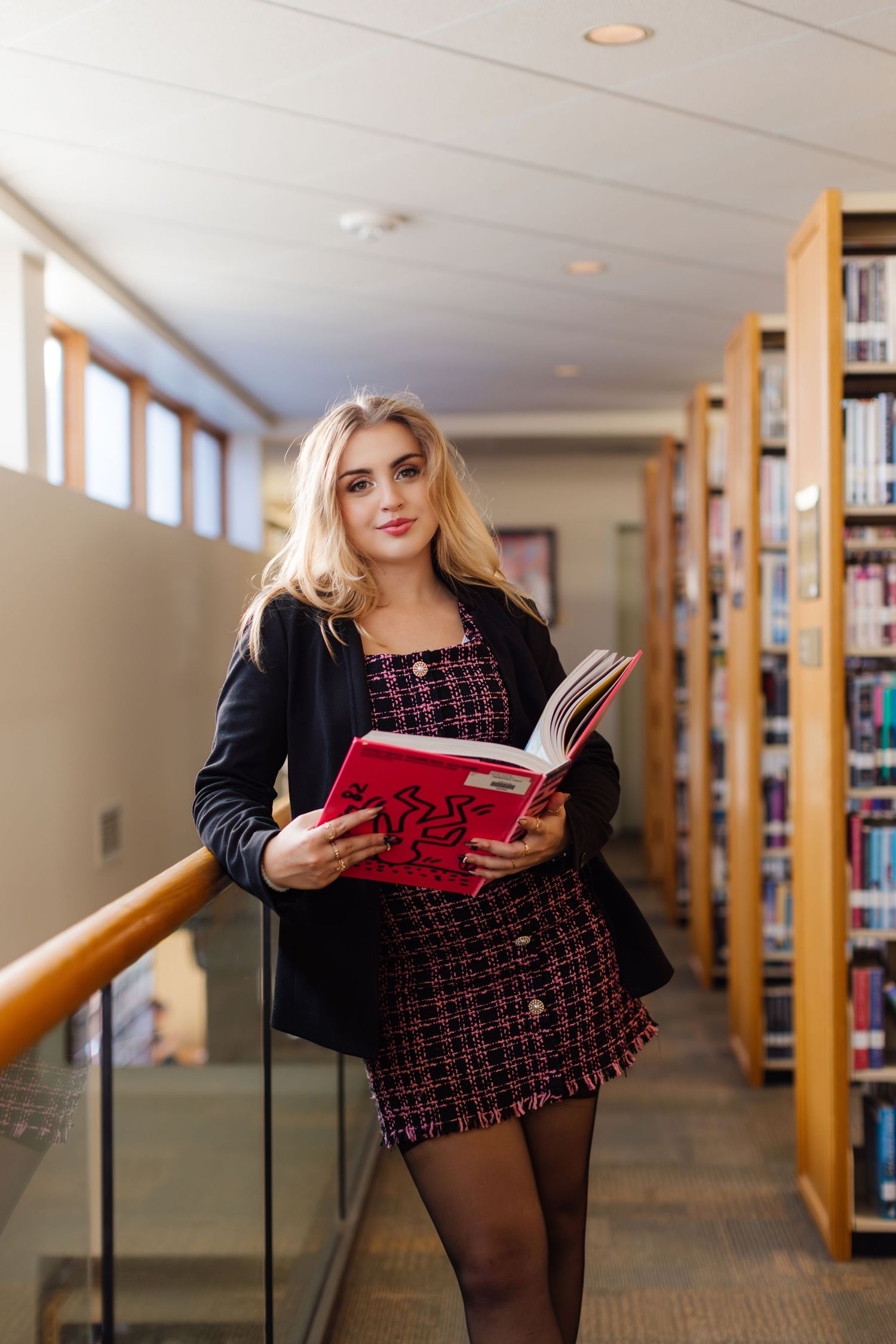 Someone browses through a book while standing in a library aisle wearing a black cardigan and patterned dress.