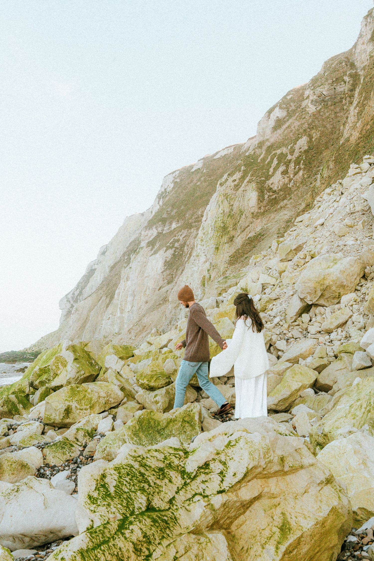 Two people climb over moss-covered rocks along a scenic white chalk cliff coastline on a misty day.