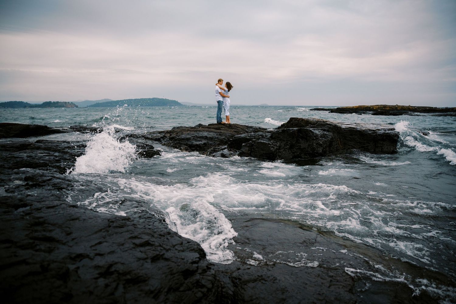 A silhouette stands on rocky coastal outcrop as waves crash dramatically against the shore during overcast weather.