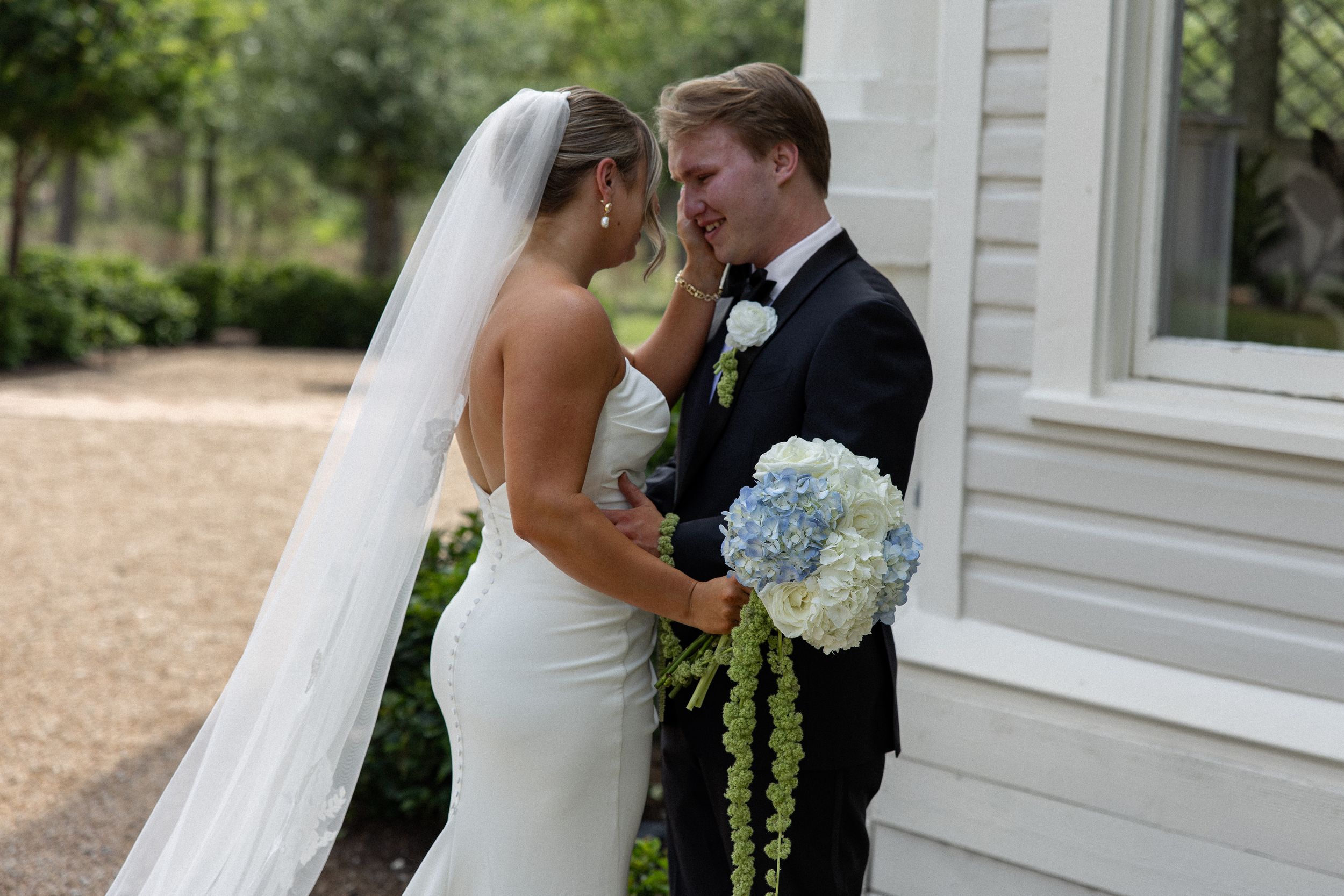 Bride in white gown and veil holds blue hydrangea bouquet with groom in black tuxedo outdoors.