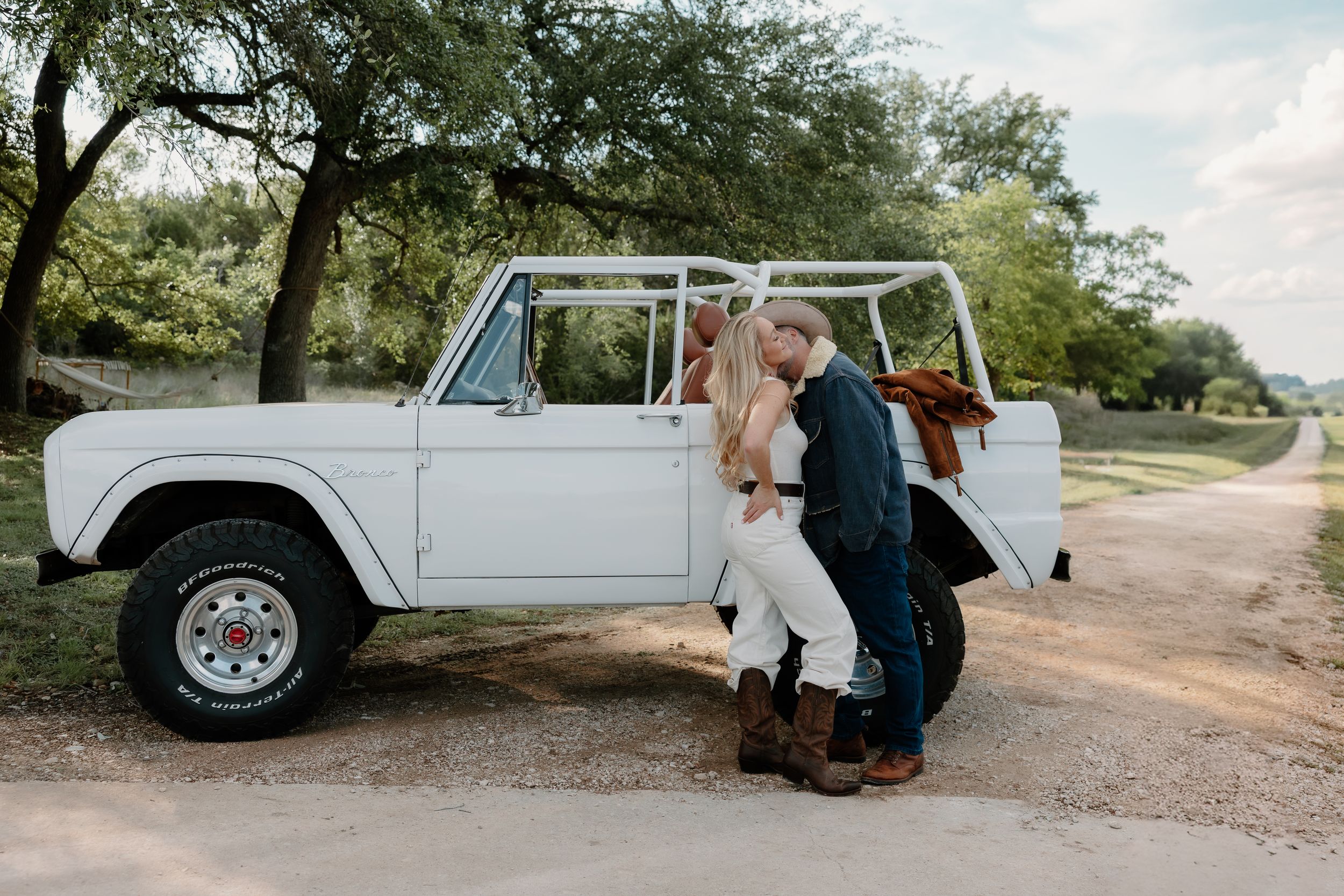 Newlywed couple posing beside vintage white Bronco SUV on rustic dirt road at outdoor wedding venue.