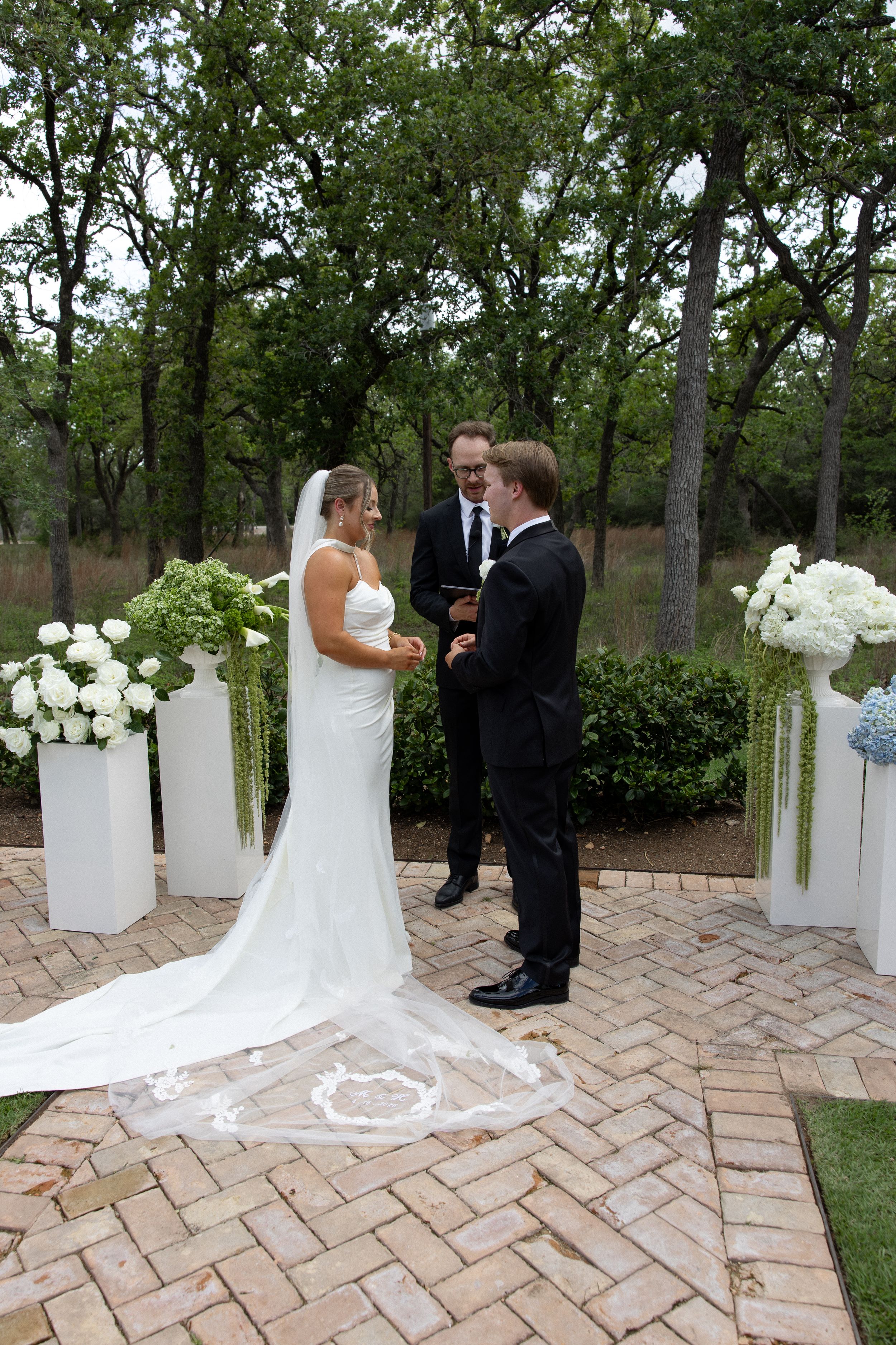 Bride and groom exchange vows outdoors on brick patio with white floral arrangements and officiant in wooded setting.