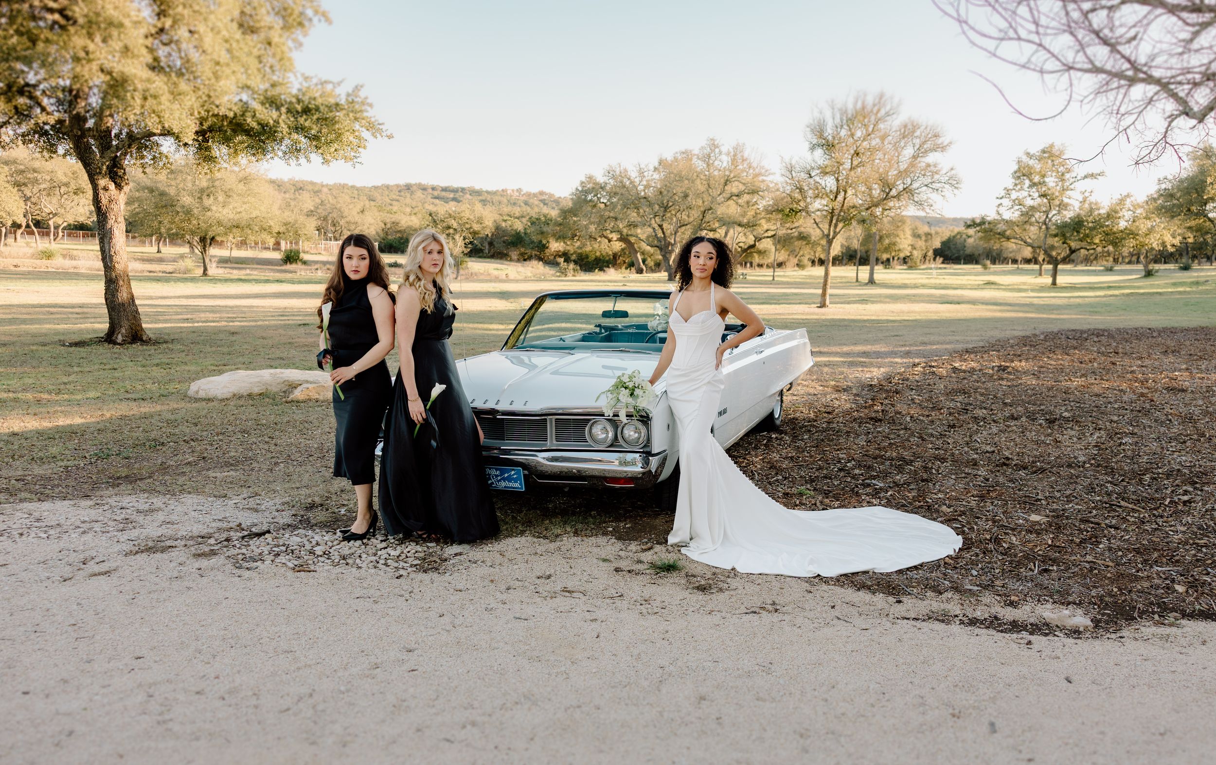 A vintage blue car serves as a backdrop for a bridal party photo shoot in a scenic outdoor park setting.