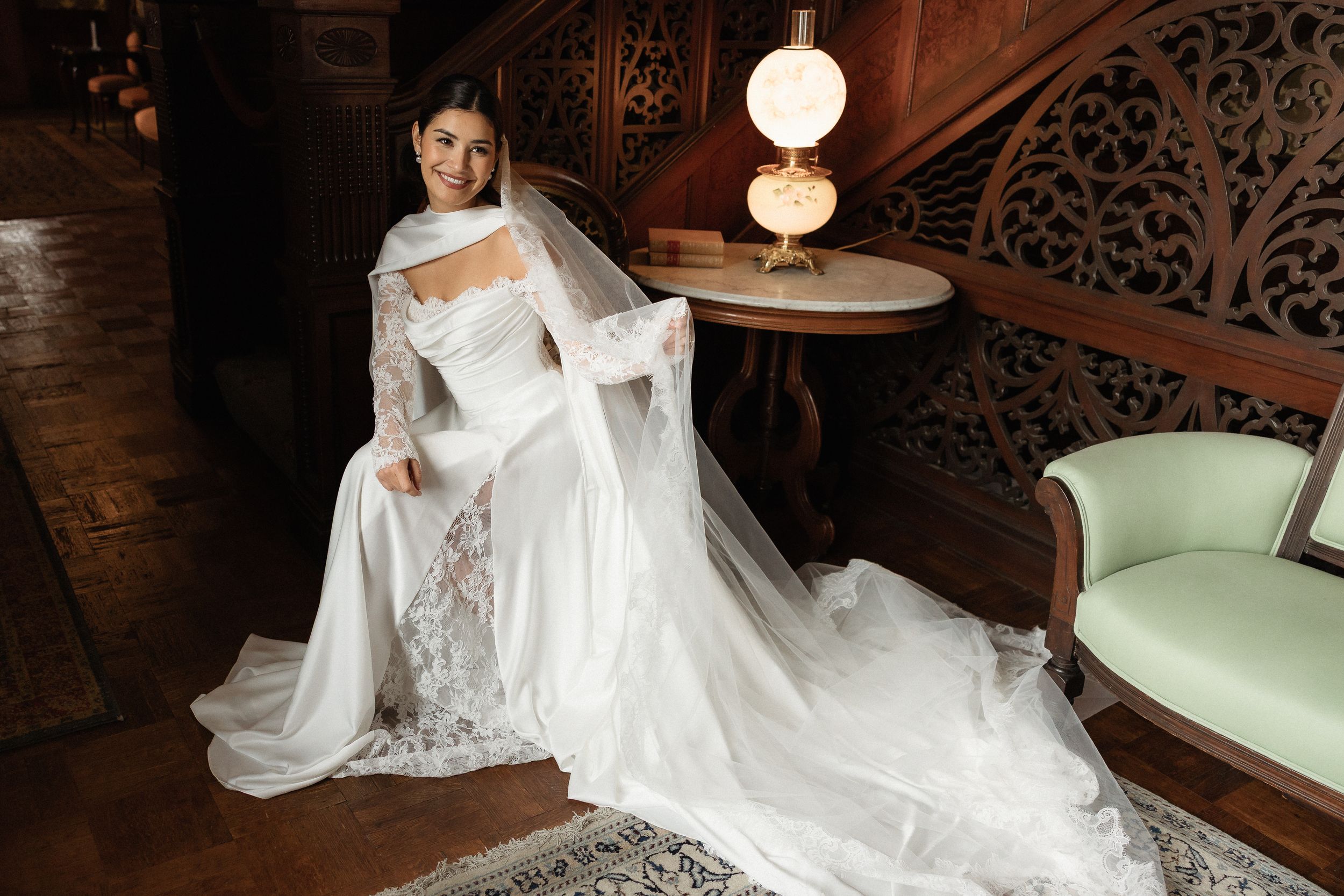 Bride in elegant white lace wedding gown with long veil seated in ornate wood-paneled room with vintage lamp.