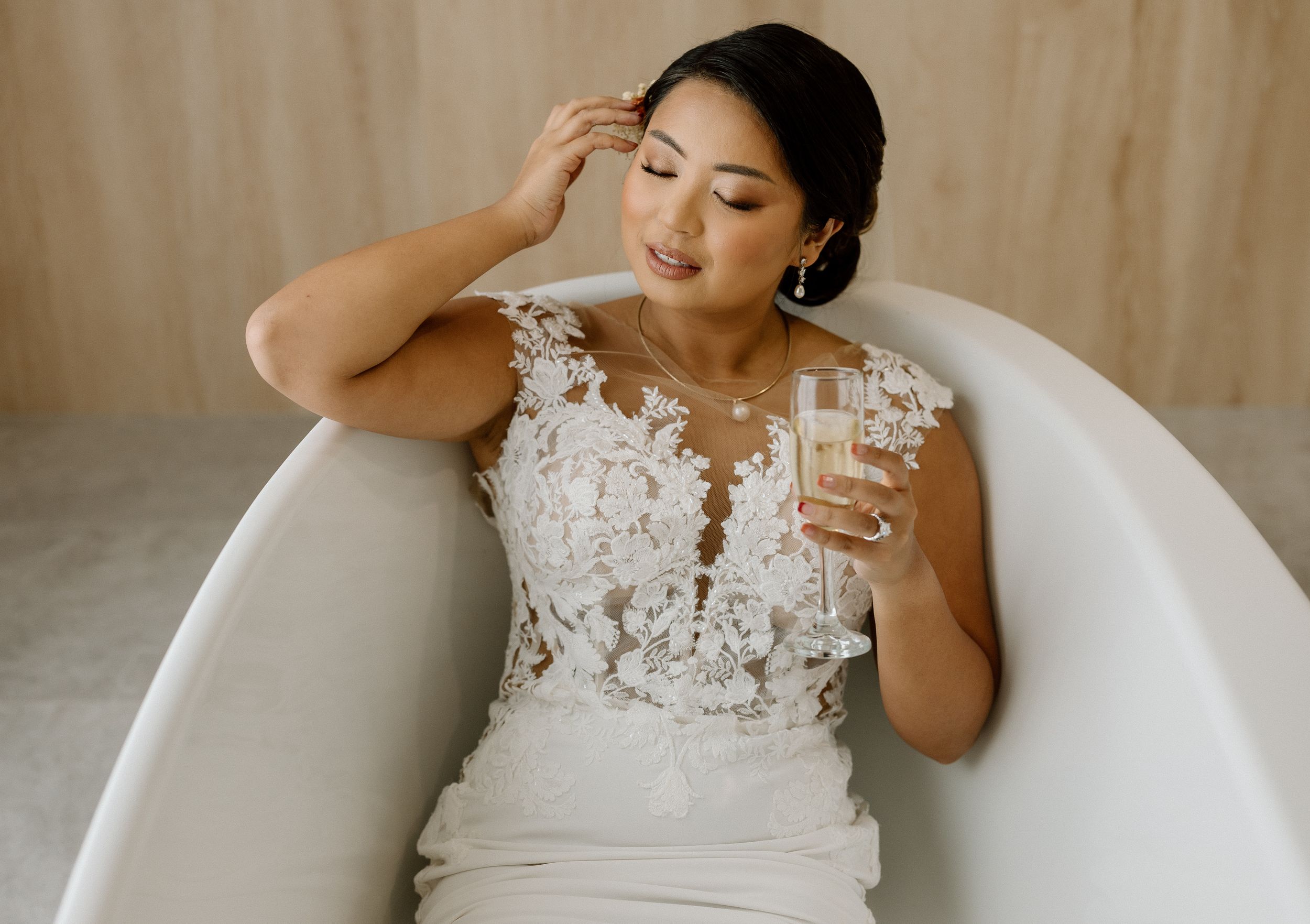 A bride in an elegant white lace wedding dress sits in a modern white bathtub holding a glass of champagne.