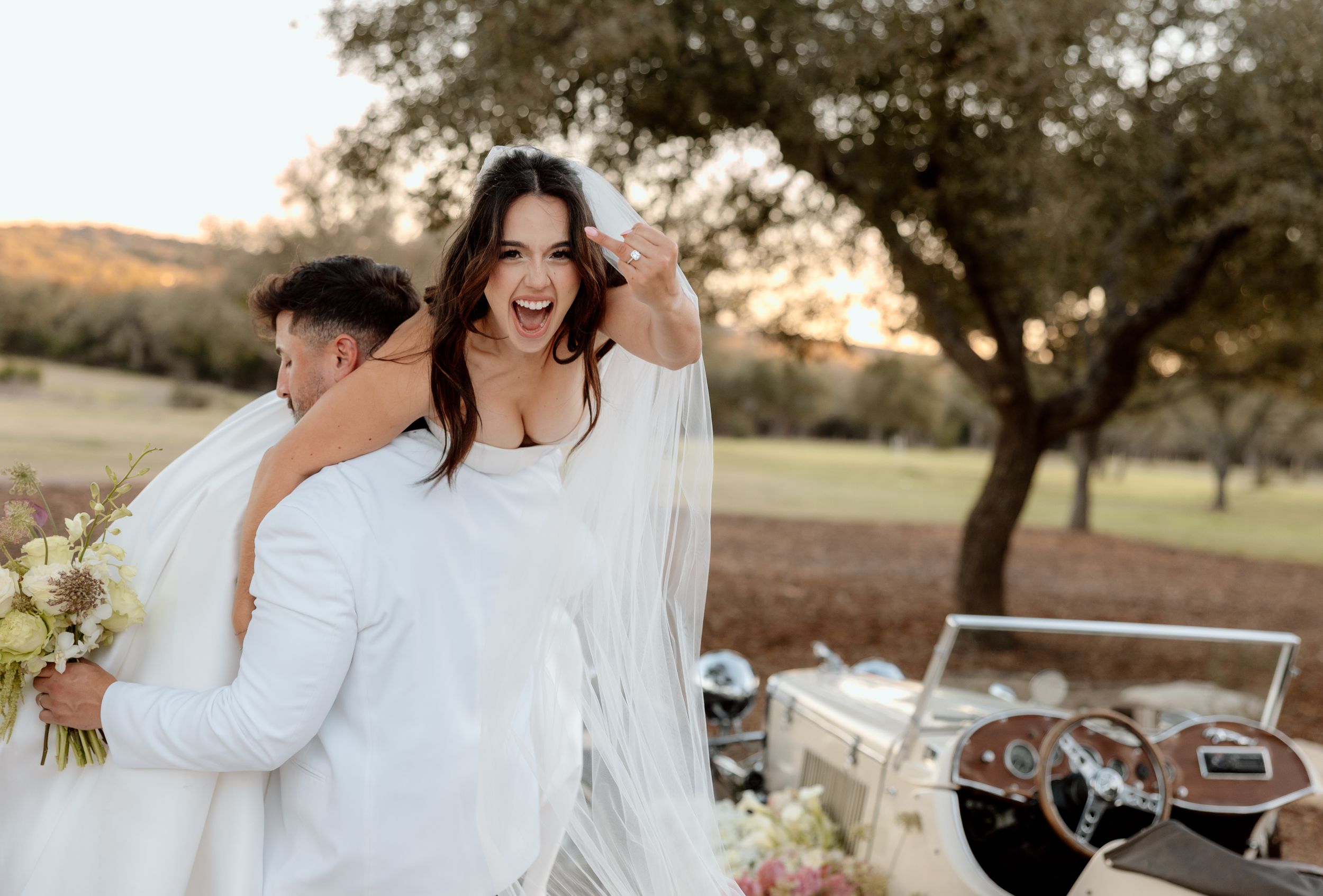 A couple in white wedding attire embrace and laugh joyfully in front of a vintage car during a scenic outdoor celebration.