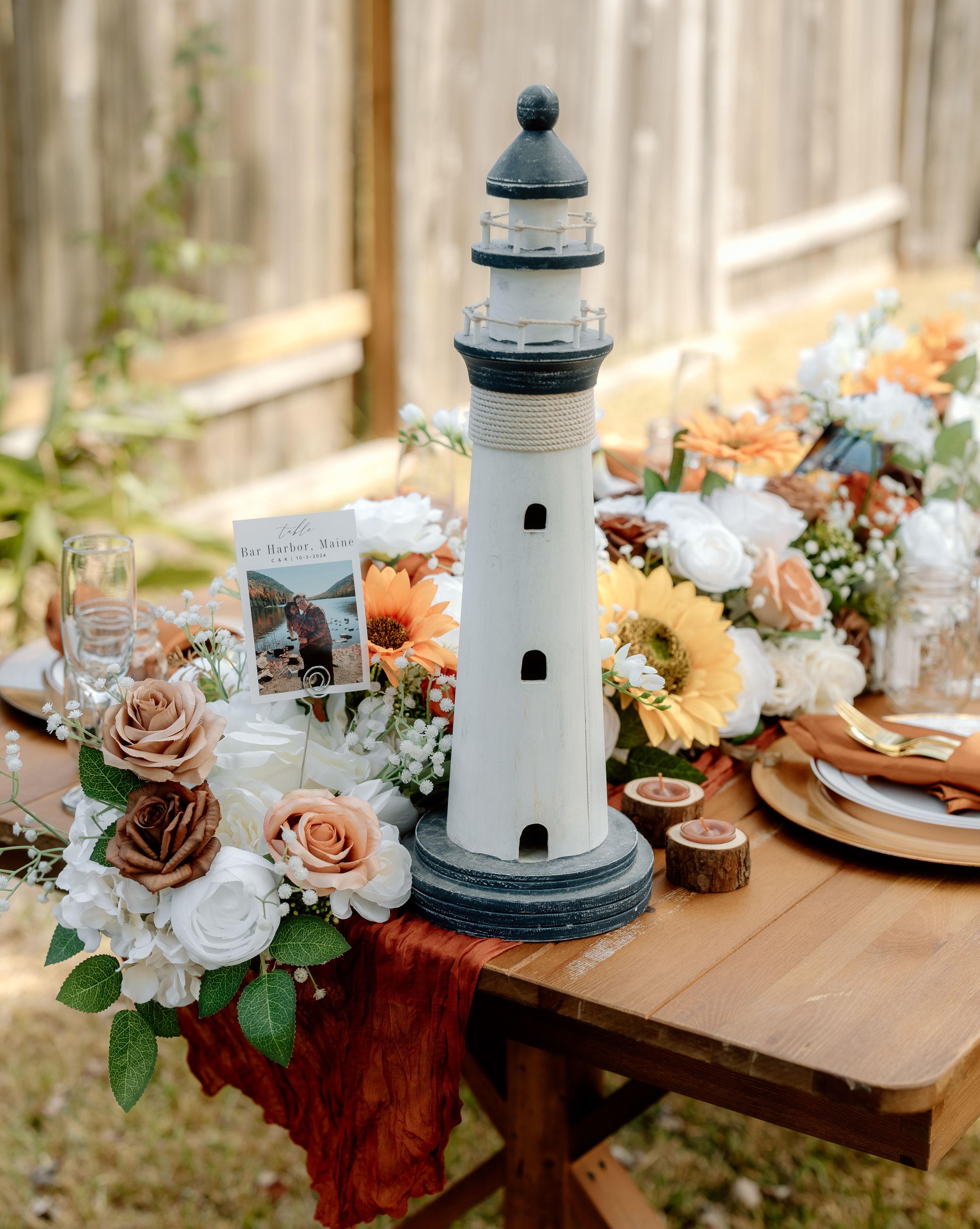 A decorative lighthouse centerpiece surrounded by floral arrangements and place settings on a rustic wooden table.