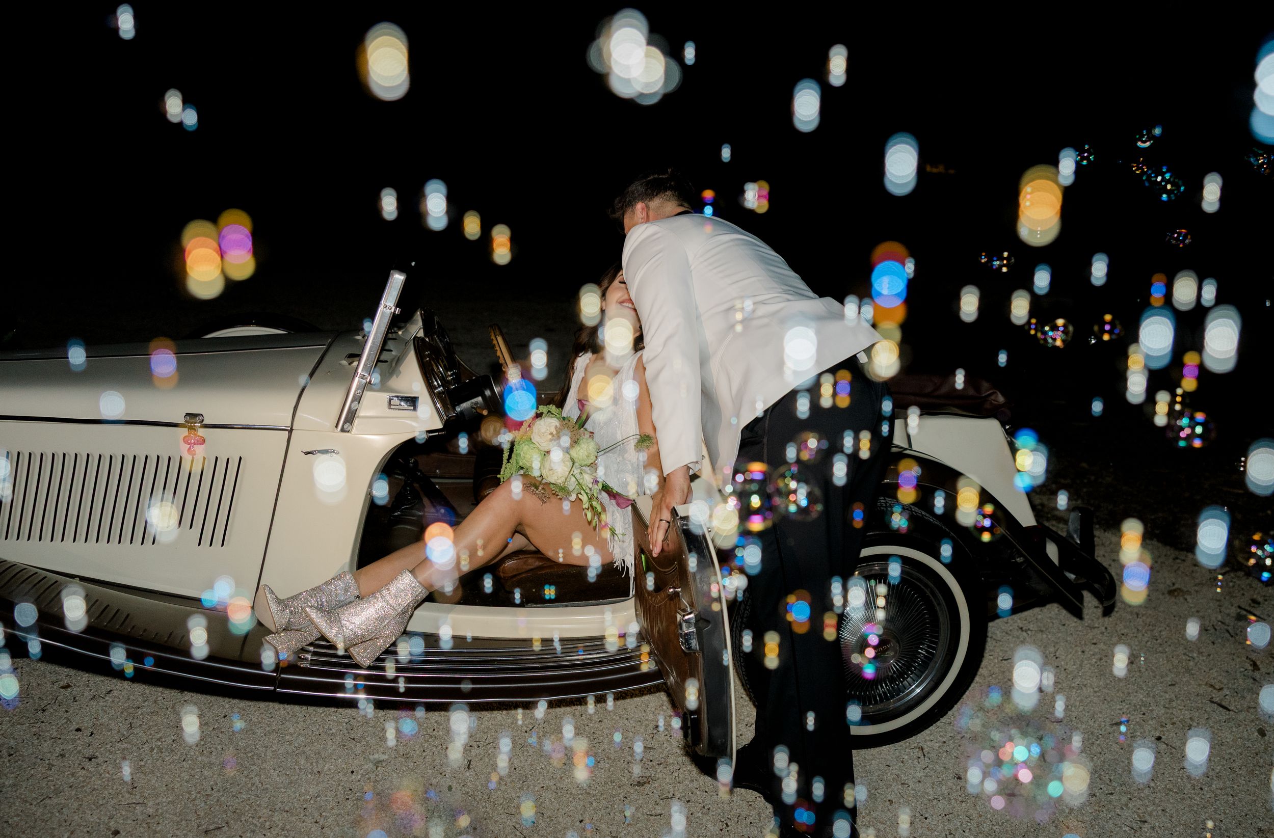 A vintage car under sparkling lights and falling water droplets captured in a dark atmospheric nighttime scene.