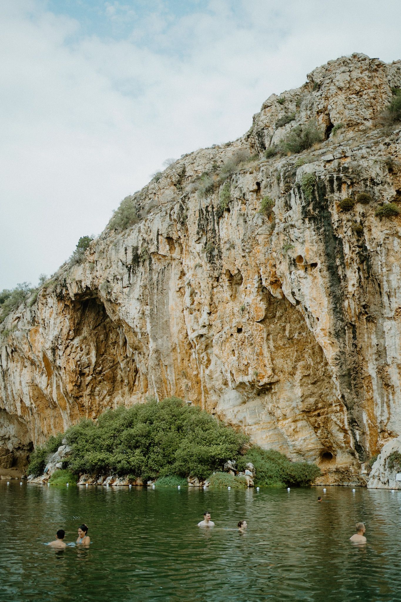 People swim in a lake surrounded by towering limestone cliffs while enjoying the natural beauty of Vouliagmeni Lake in Greece.