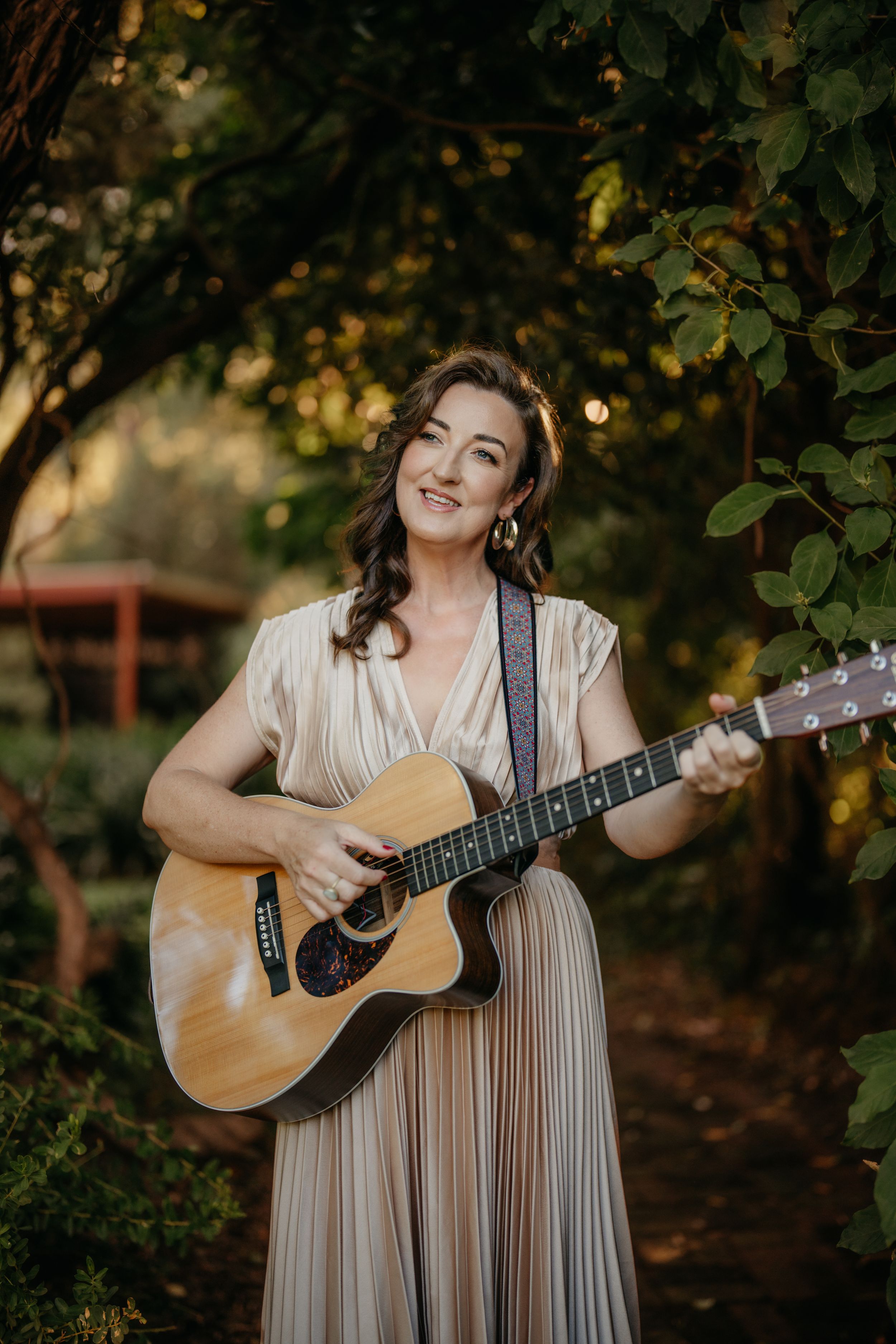 Smiling brunette woman in a striped dress playing acoustic guitar surrounded by lush green foliage.