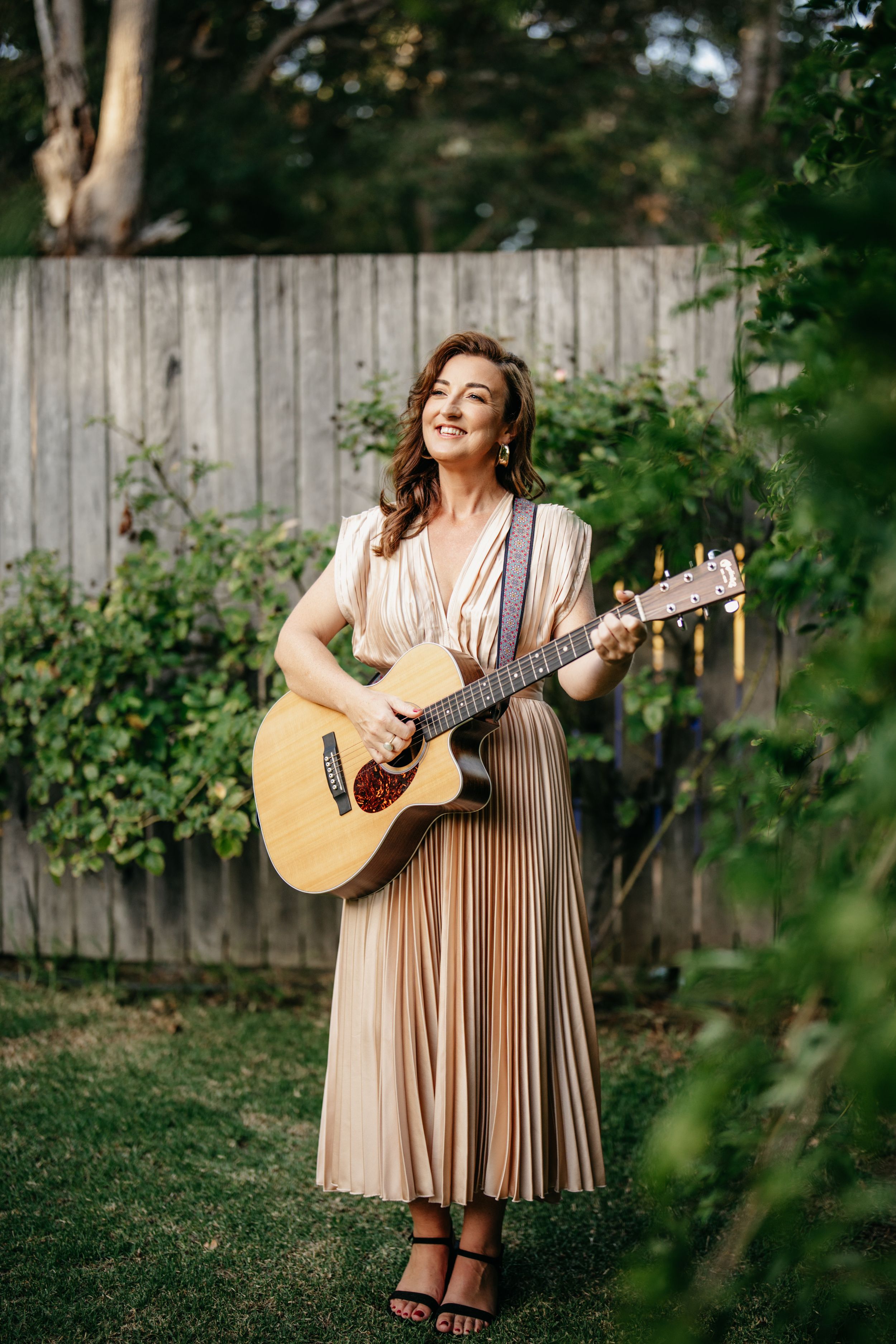 Smiling woman in a beige pleated dress playing acoustic guitar while standing in a garden outdoors.