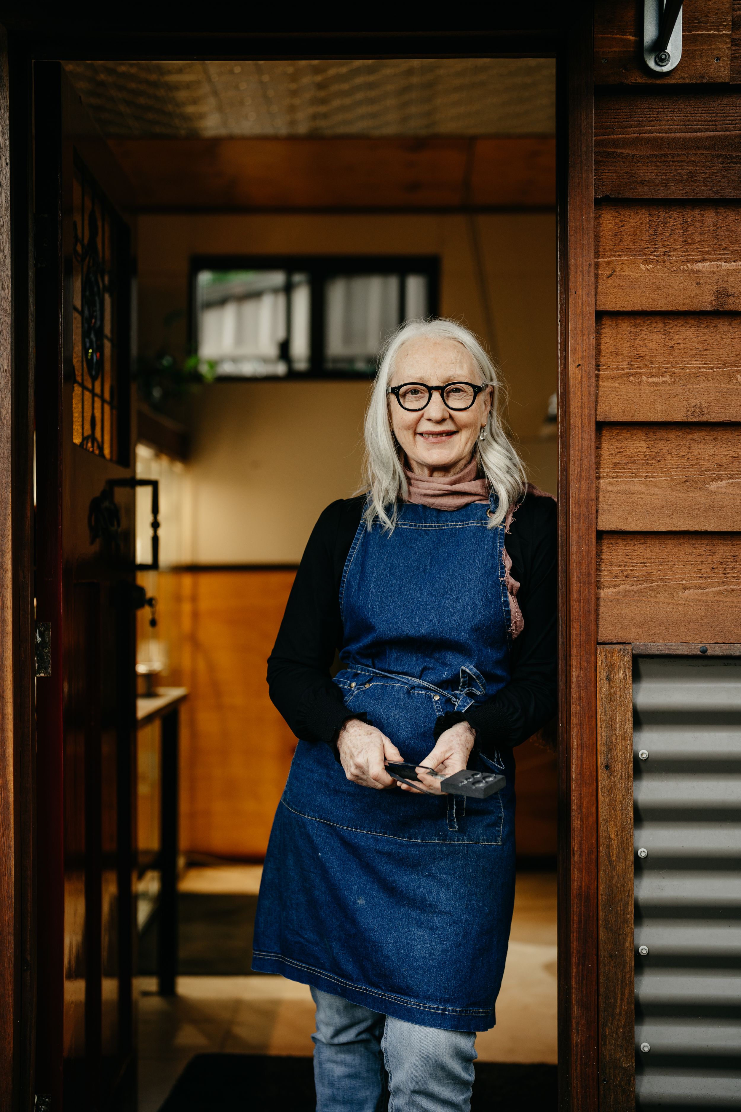 Smiling senior woman with grey hair and glasses wearing a denim apron standing in a wooden doorway.