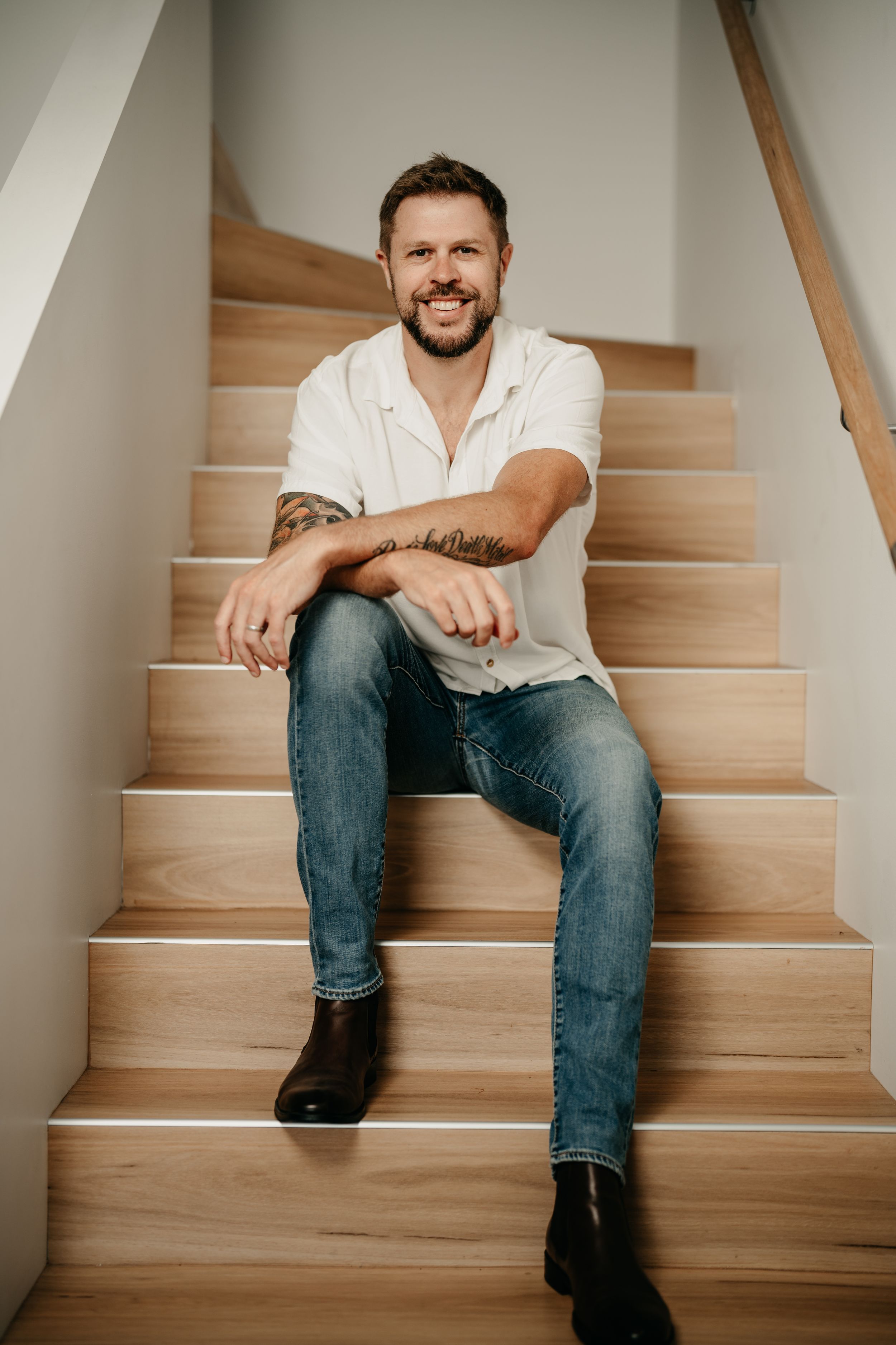 Smiling man with tattoos sitting on wooden stairs wearing white shirt and jeans.