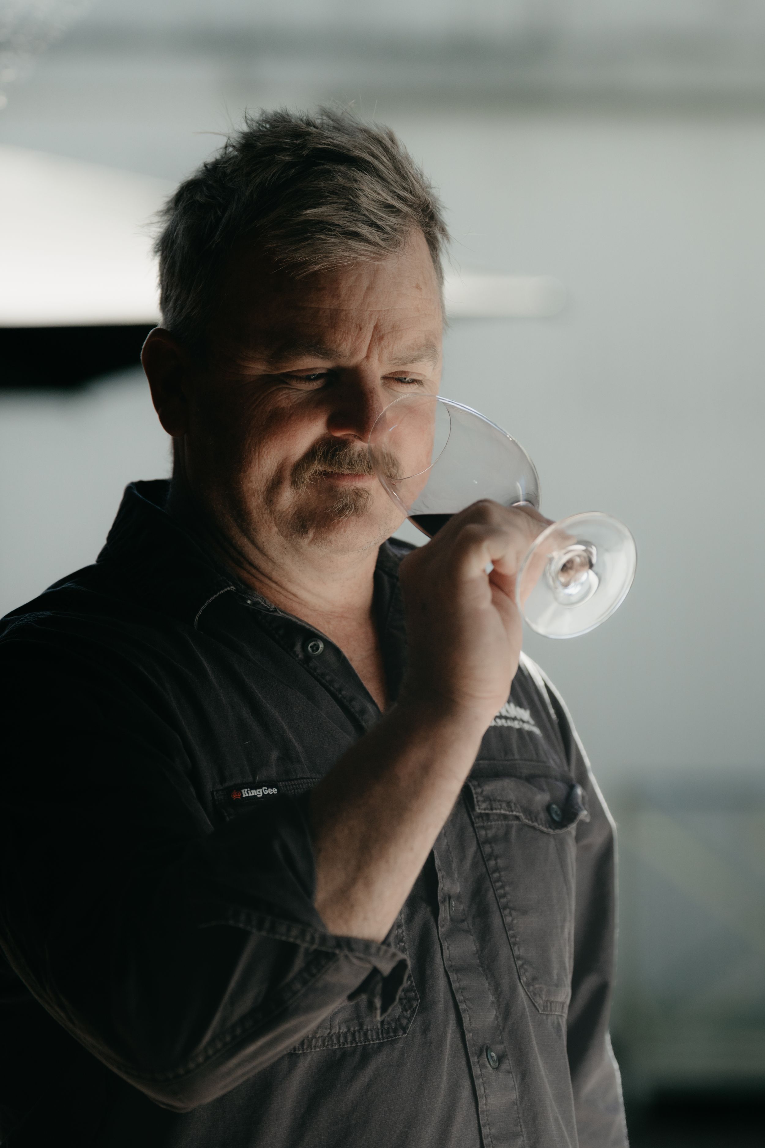 Middle-aged man with a mustache smelling a wine glass in a dimly lit room wearing a dark shirt.