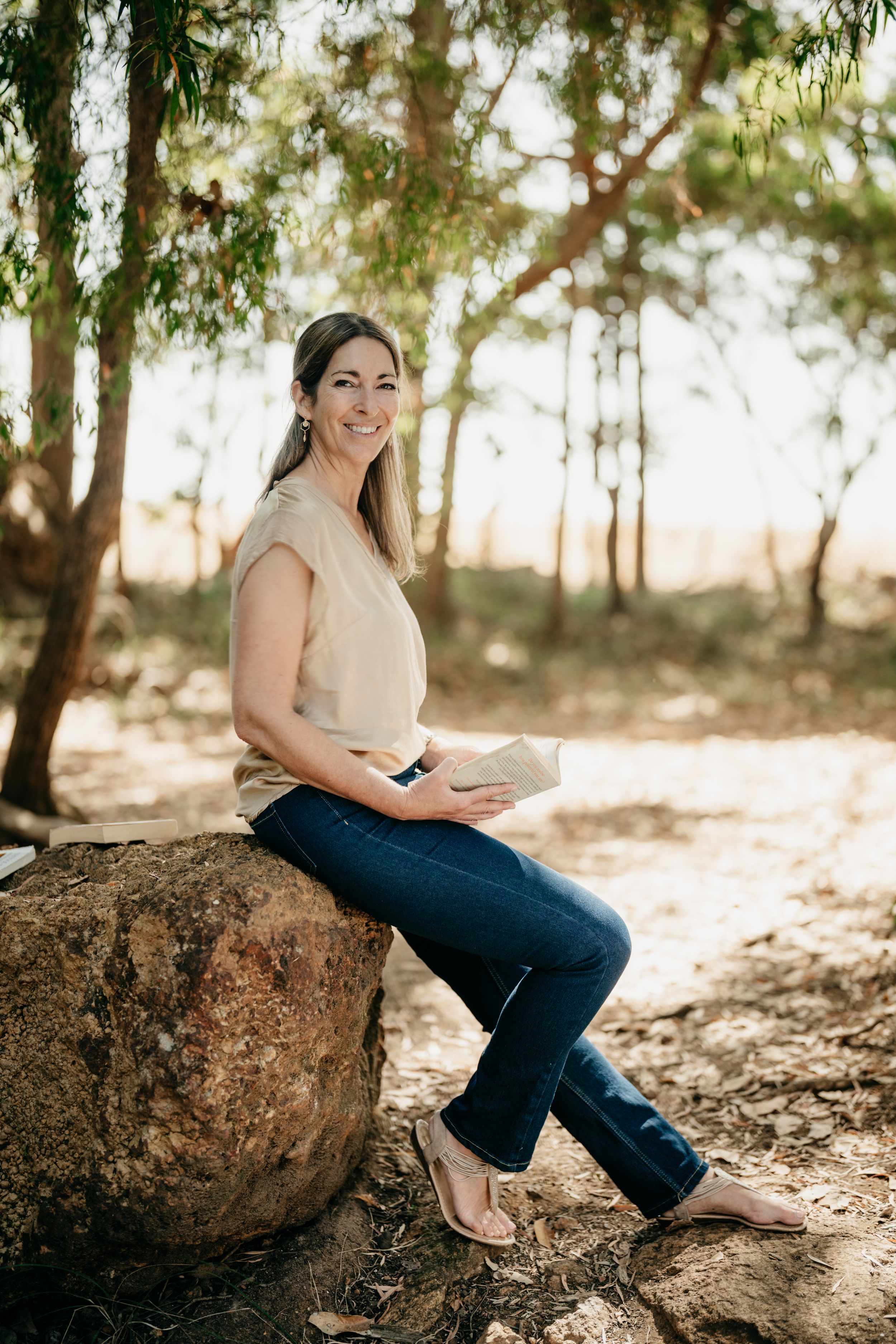 Smiling woman in jeans and beige top sitting on a rock holding a book in a sunlit eucalyptus forest.