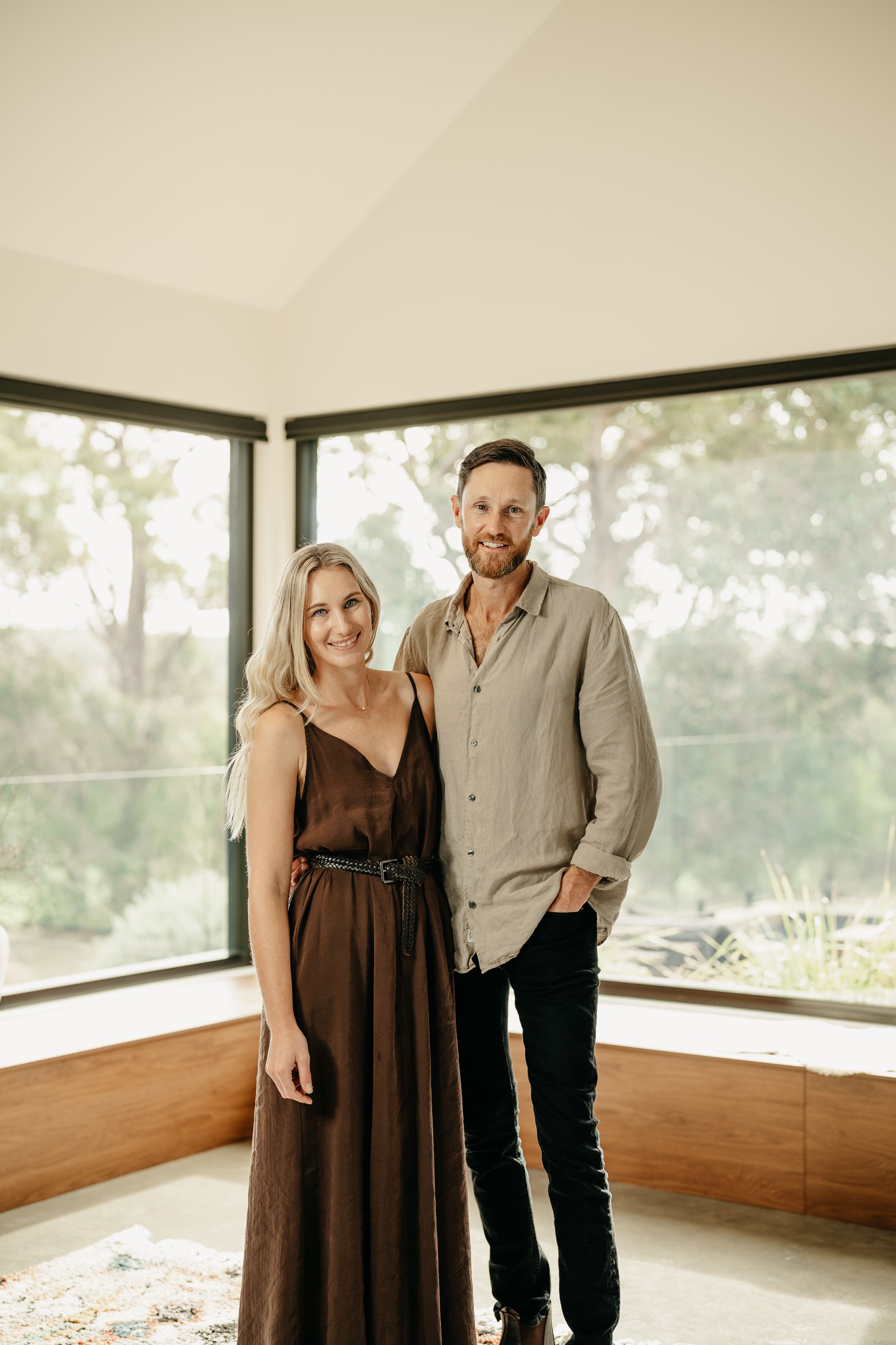 Happy couple standing together in a bright sunlit room with large windows overlooking green trees.