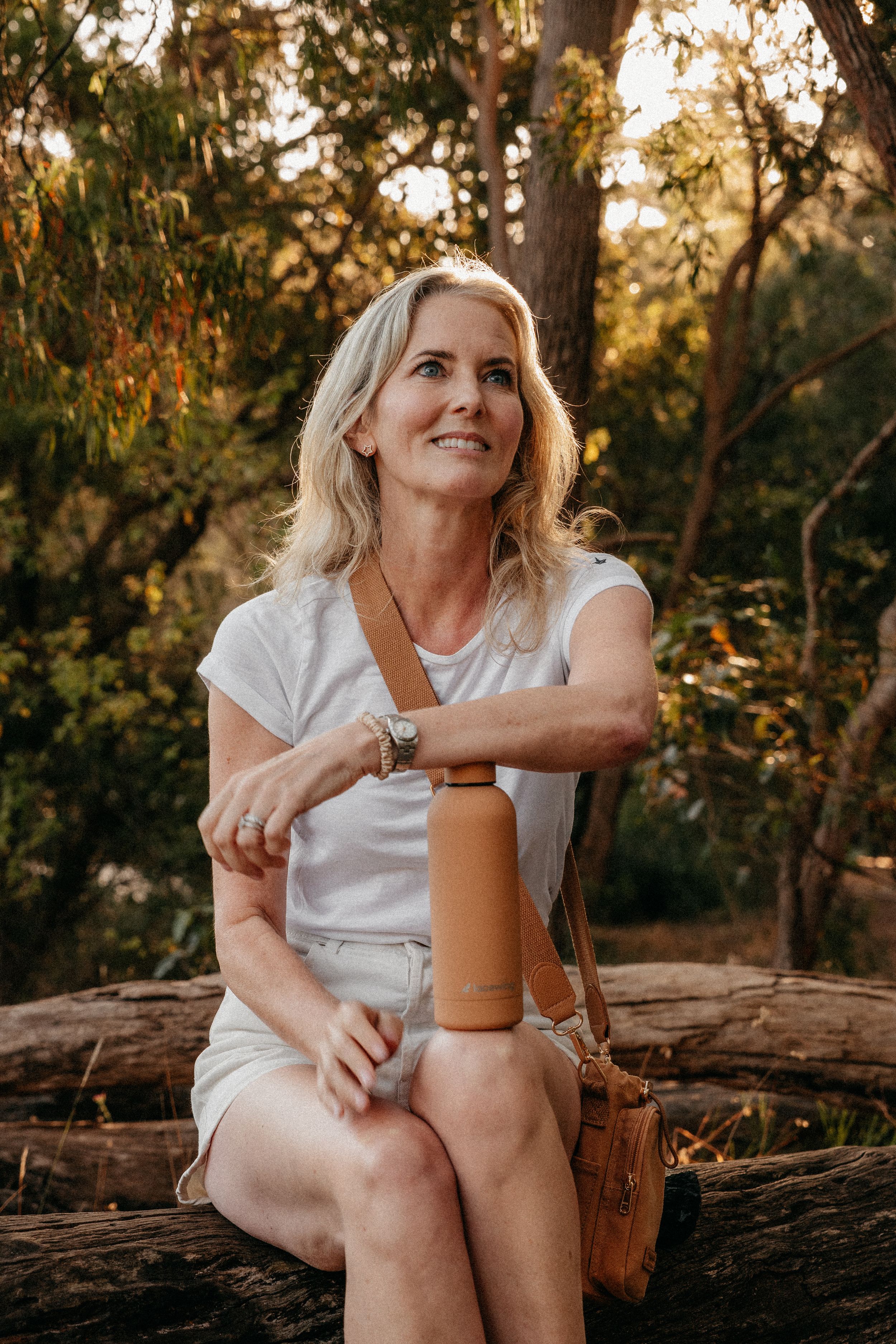 Blonde woman sitting on a log in nature holding a tan water bottle, smiling in golden sunlight.