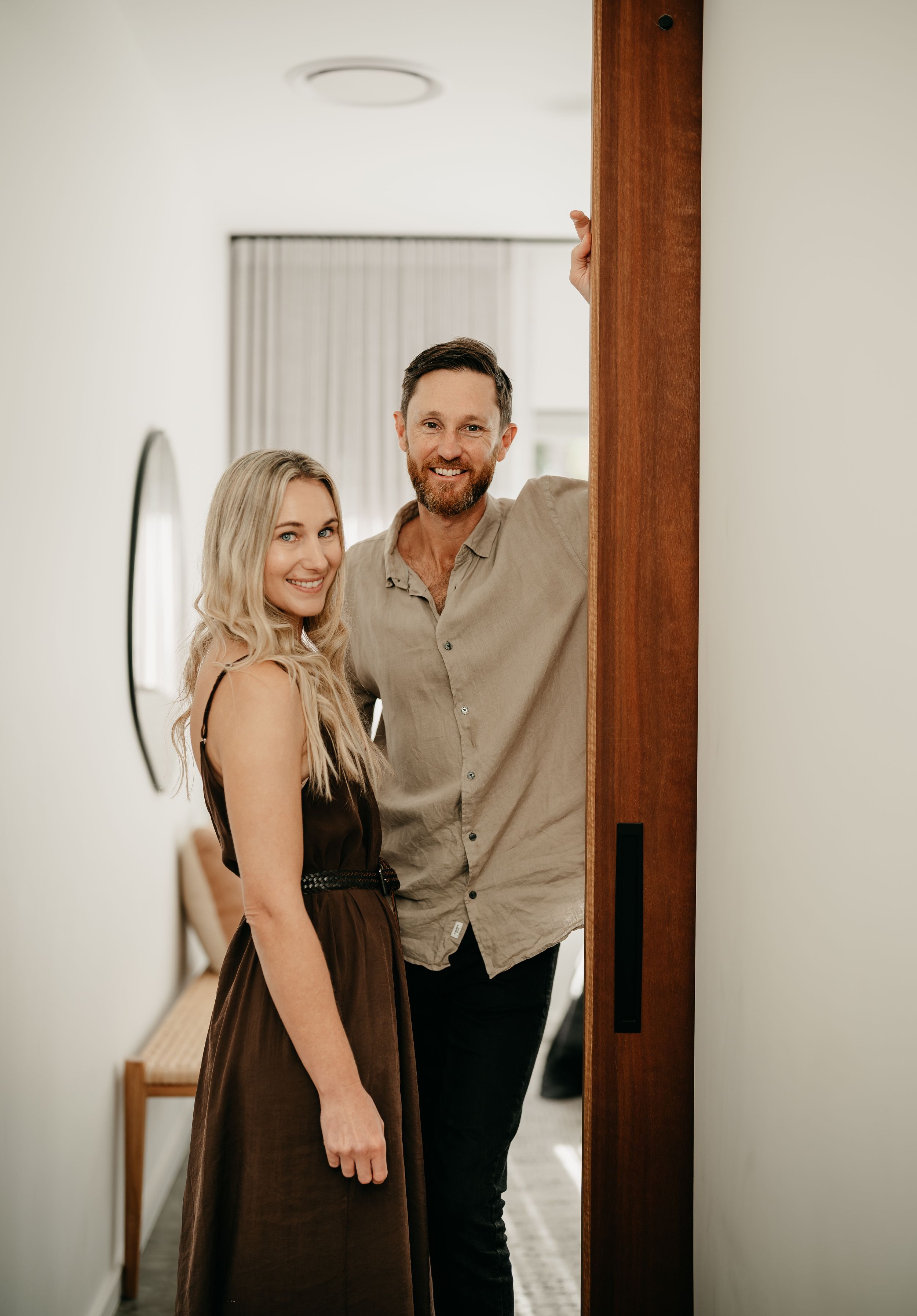 Smiling couple posing together in a modern home doorway, woman in brown dress and man in grey shirt.